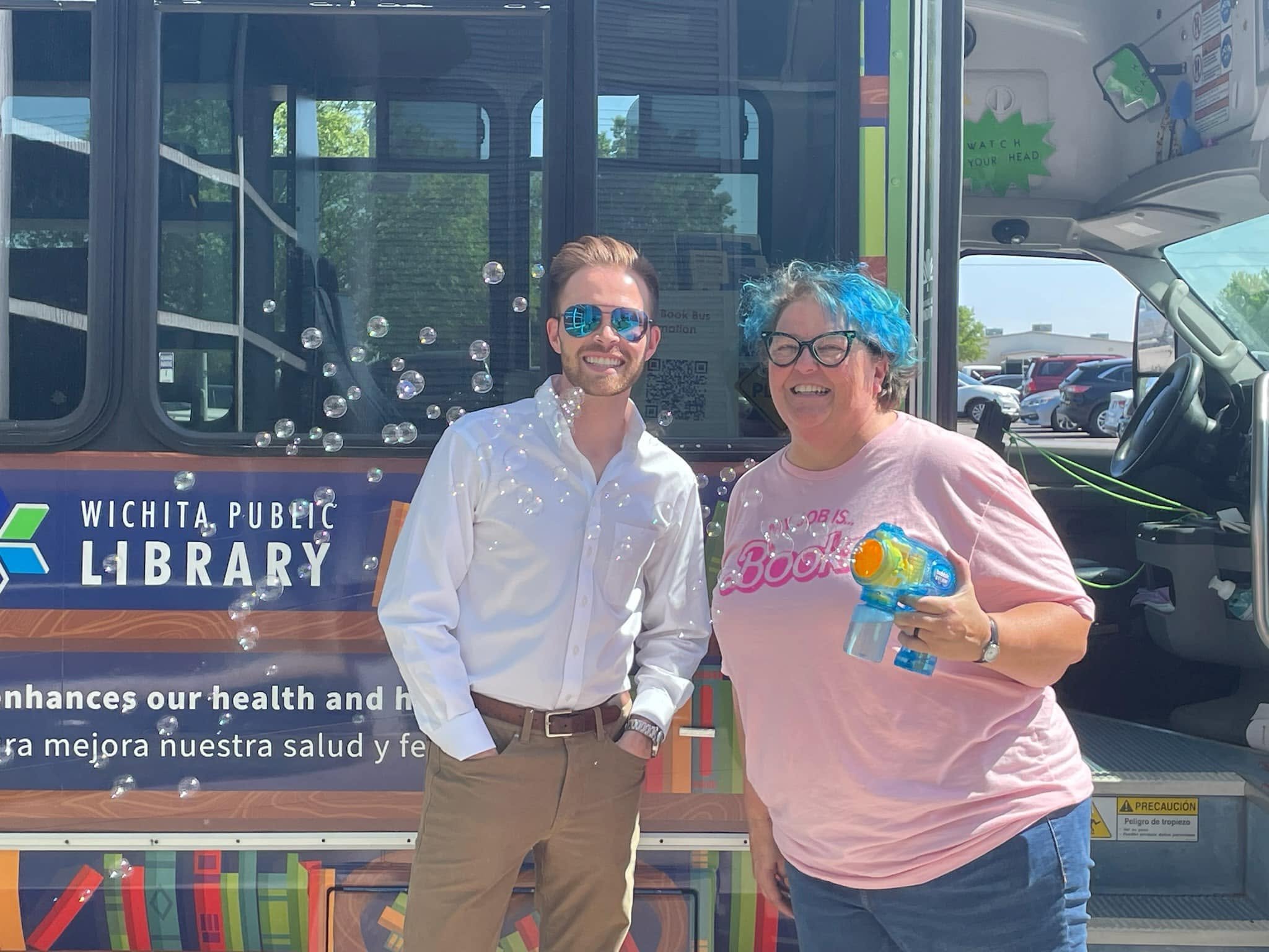 Two people smiling in front of a bus with the logo for the Wichita Public Library. One person is wearing sunglasses and a white shirt, the other has colorful blue hair and glasses, holding a water gun.