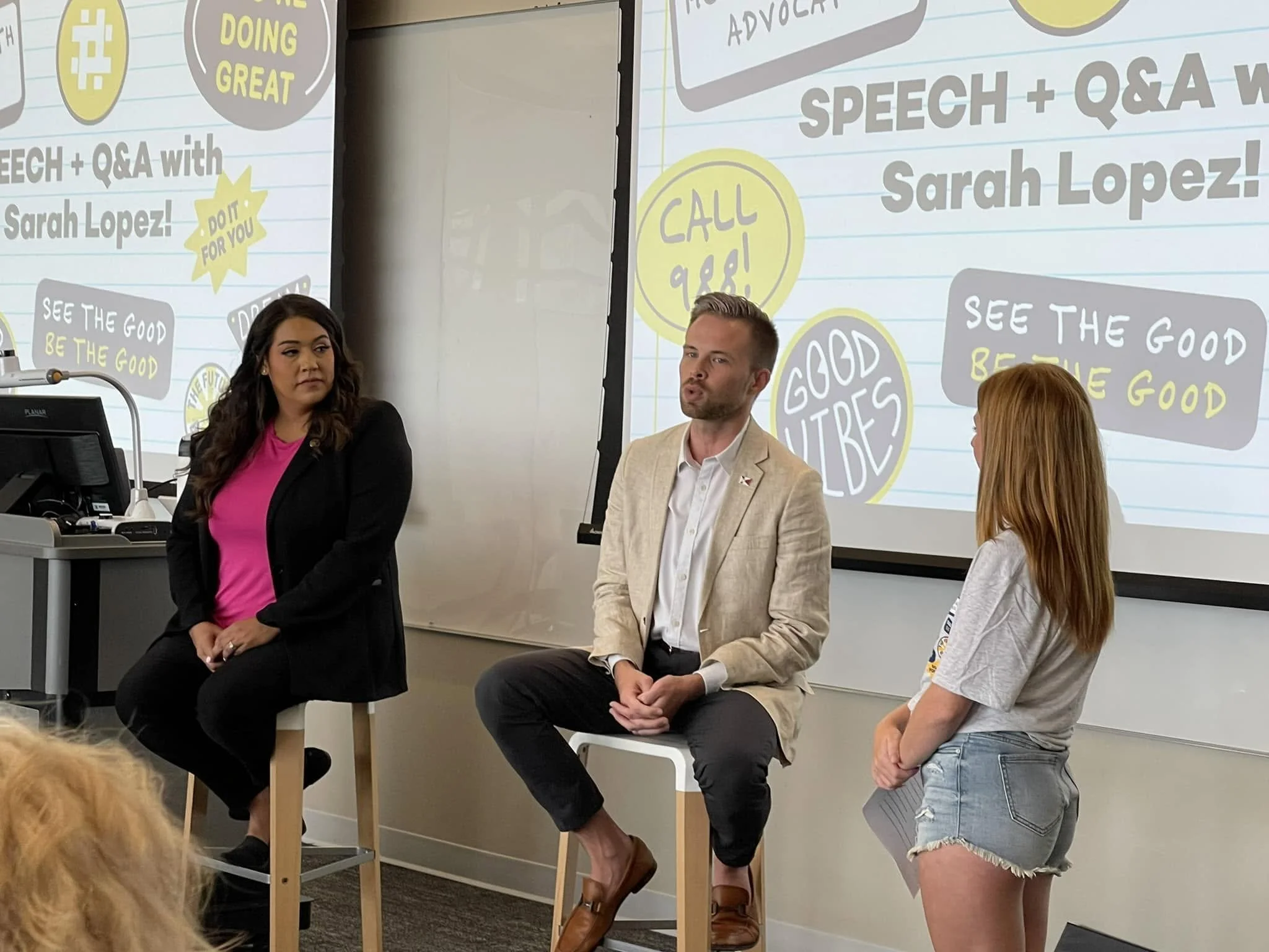 Three panelists seated in front of a large screen displaying a presentation about speech and Q&A, with animated text bubbles and icons, in a classroom or conference room. Two female and one male panelist are present, with the female on the left weari