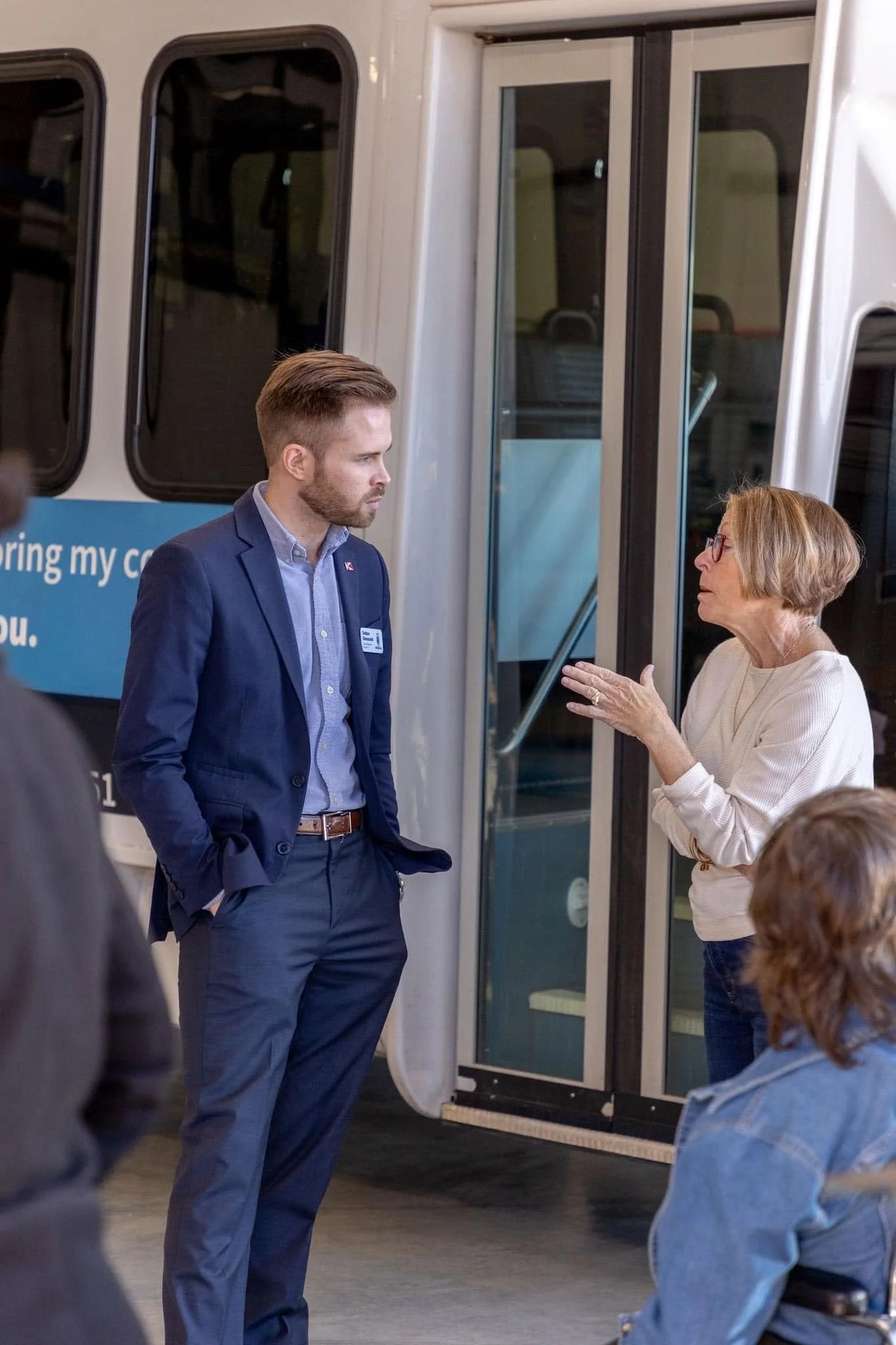 A man in a navy suit and a woman wearing glasses and a beige sweater are talking in front of a bus, with other people partially visible in the foreground.