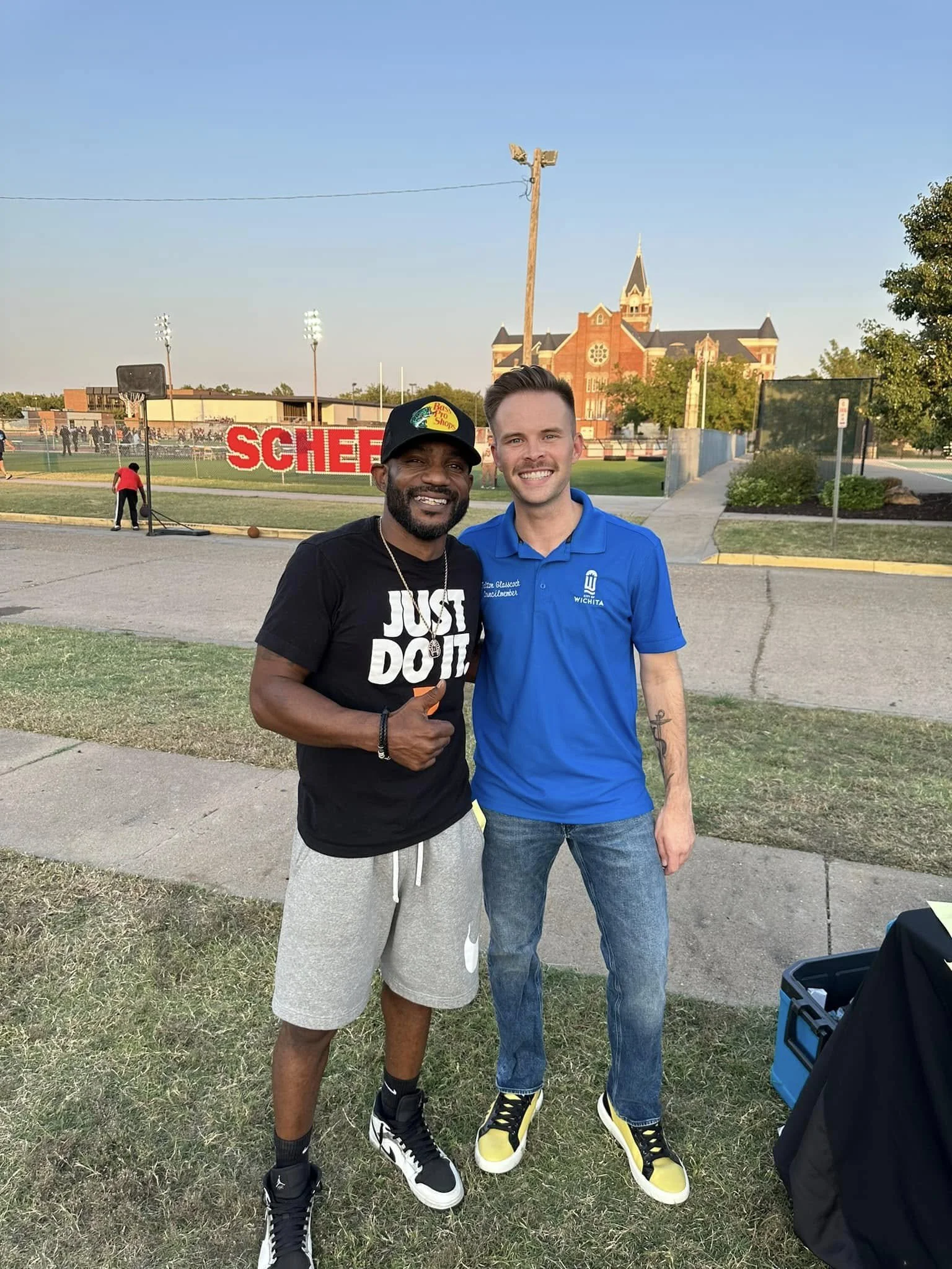 Two men smiling and standing outdoors on a grassy area, with a school and a church in the background. One man is wearing a black T-shirt with 'JUST DO IT' and a black cap, the other is in a blue polo shirt and jeans.