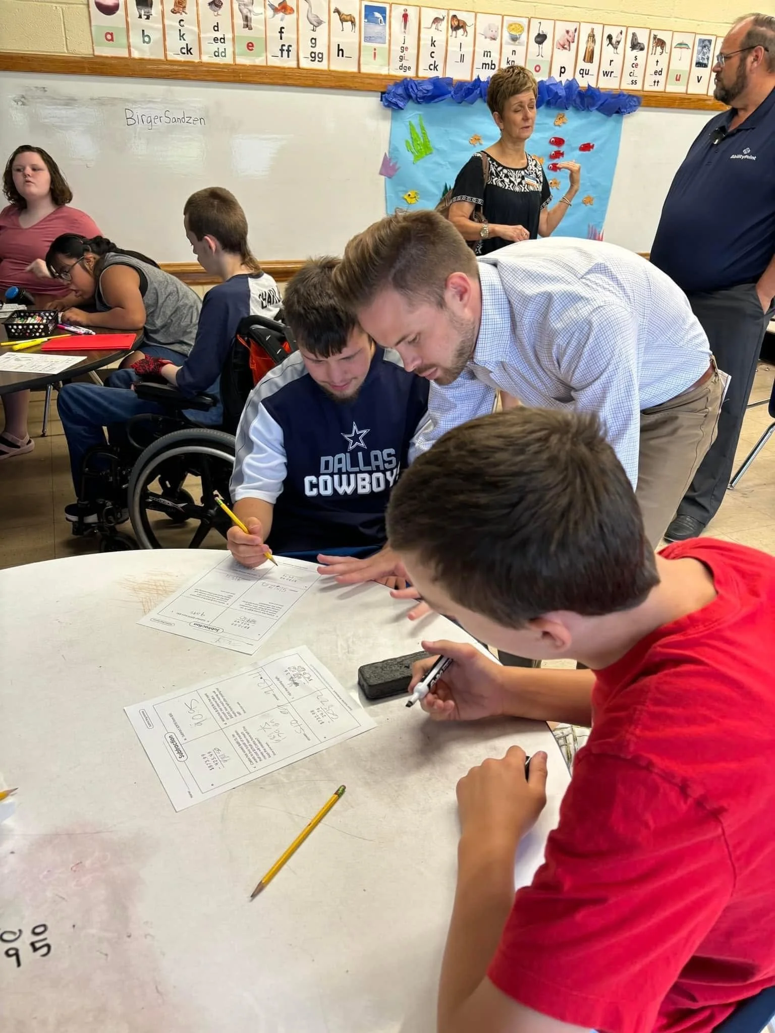 A classroom scene with students working at tables, a teacher explaining near a decorated board, whiteboard with writing, and educational posters above.