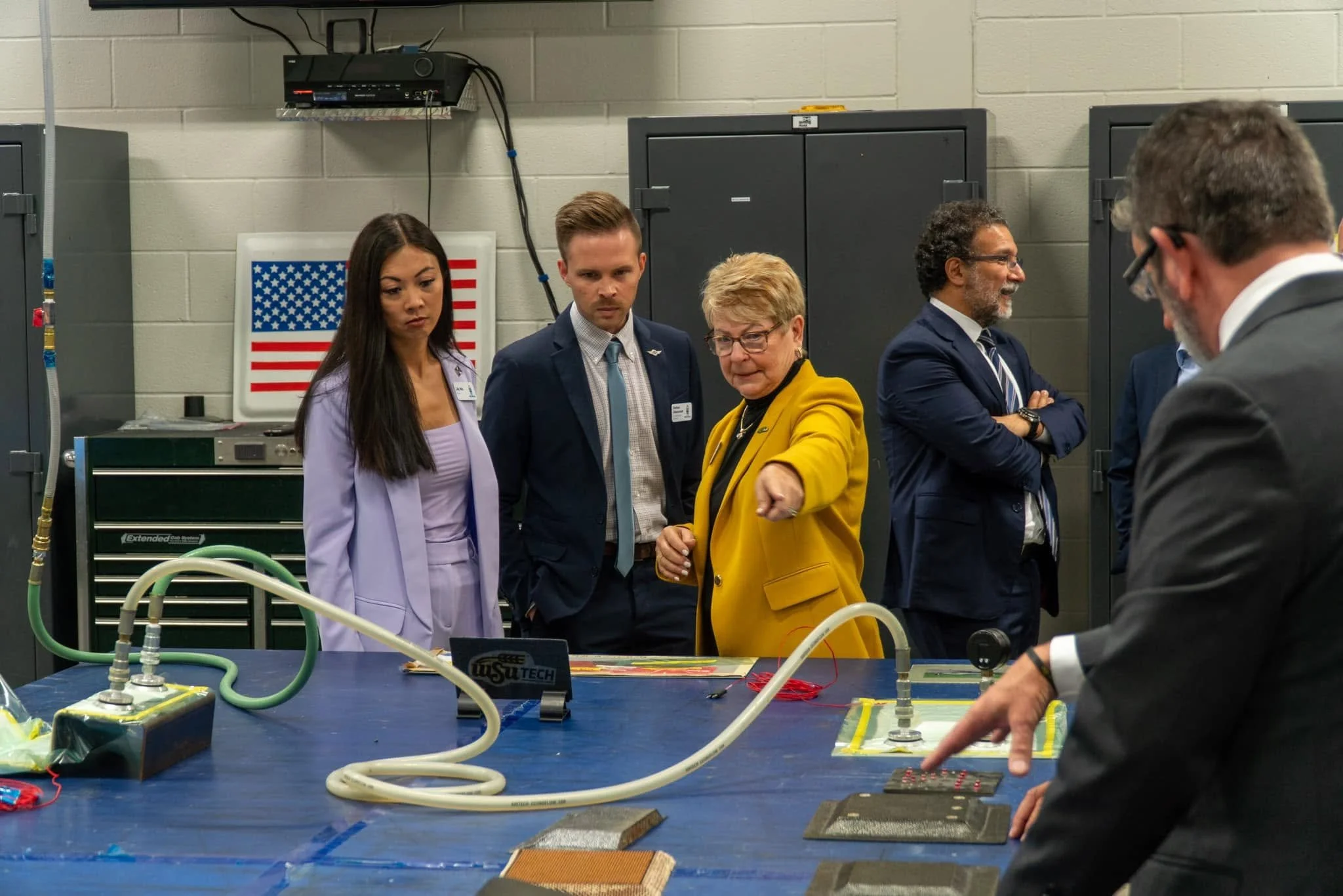 Group of people in a laboratory, observing a science experiment or demonstration at a blue worktable, with equipment and wires visible, and American flag in the background.