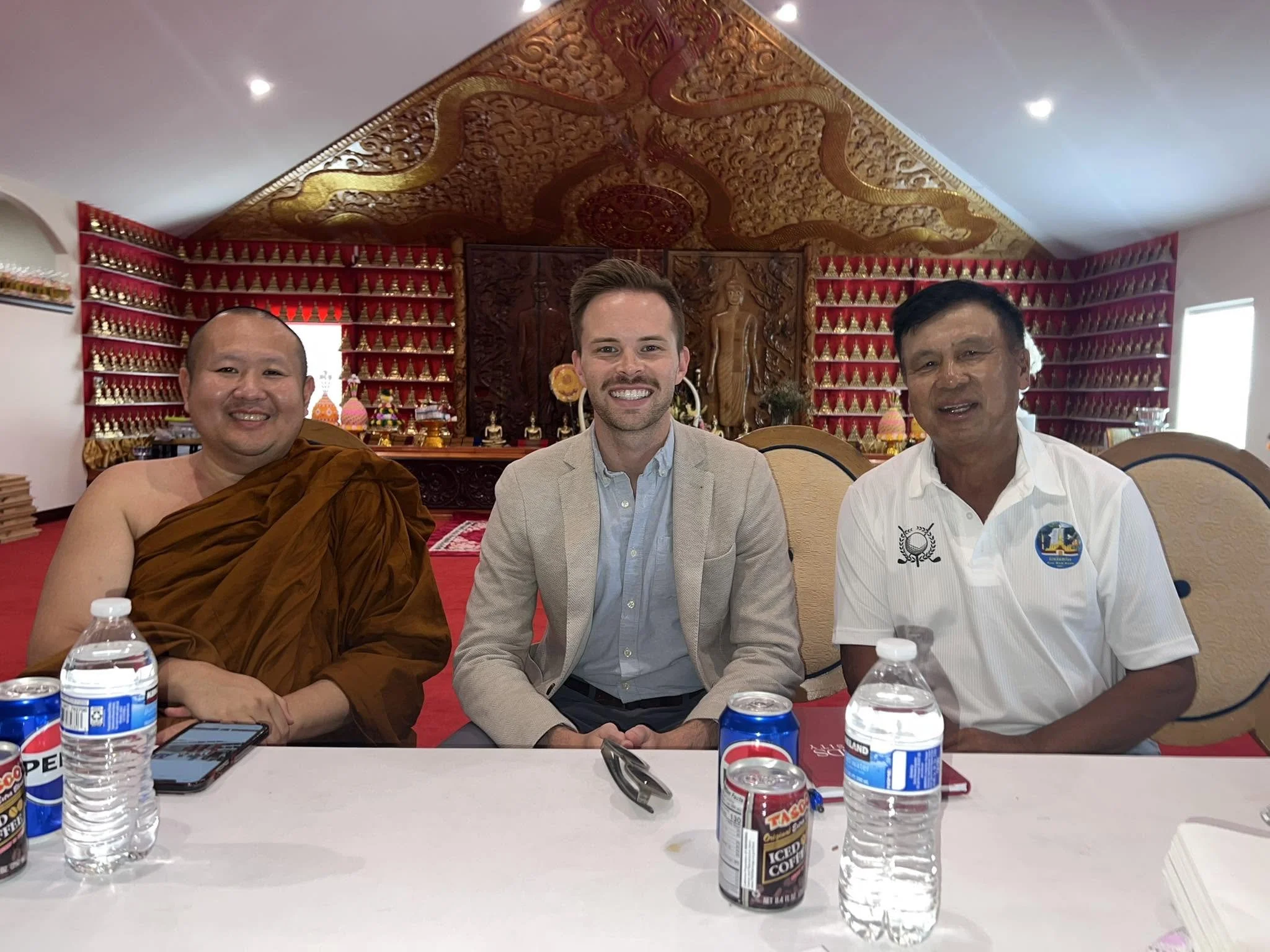 Three men sitting at a table during a meal or meeting, with bottled water and soda cans in front of them. The background features ornate woodwork and red shelves filled with small Buddha statues.