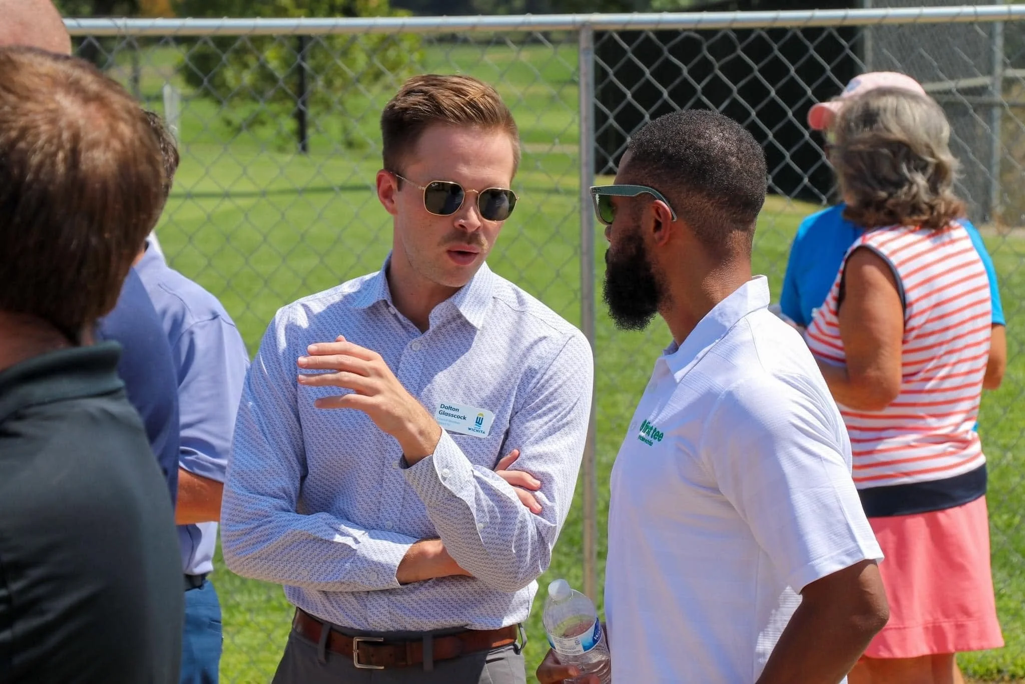 Group of people engaged in conversation outdoors near a fence, with trees and grass in the background.