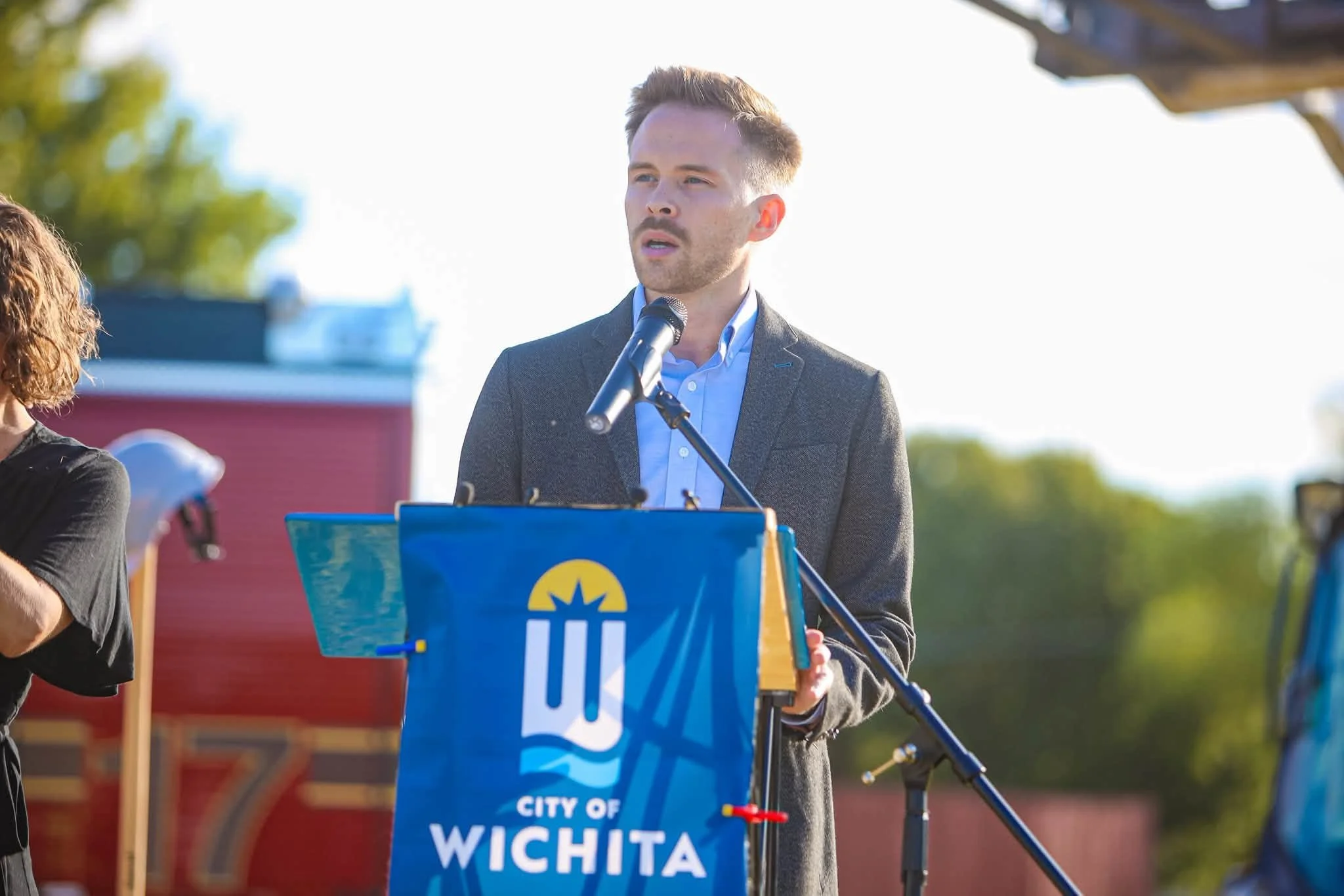 A man in a gray suit jacket and light blue shirt speaking at a podium with a microphone during an outdoor event in the daytime. The podium has a sign with the words 'City of Wichita' visible. There are trees and a red building in the background.