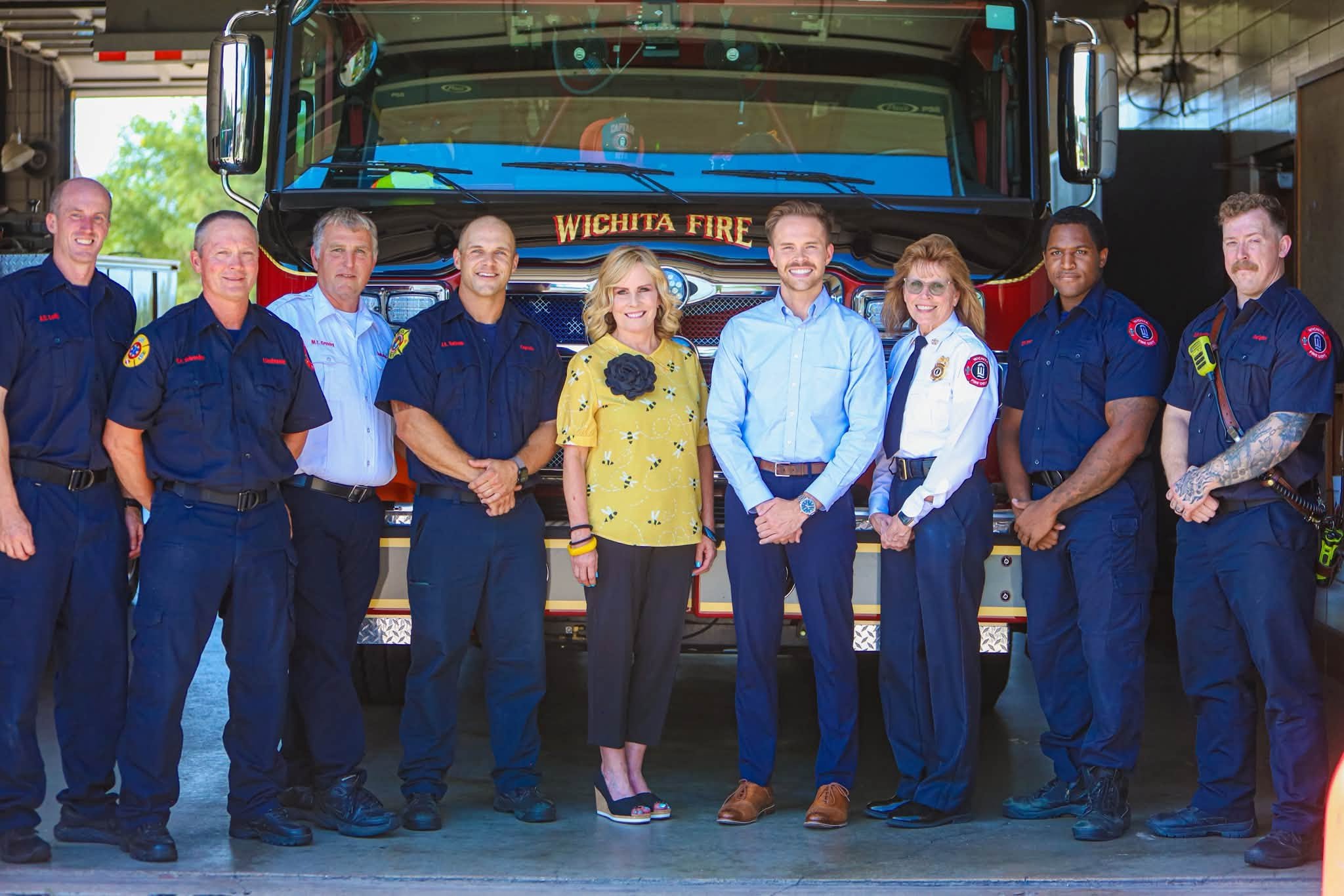 A group of nine people, including firefighters and women, standing in front of a fire truck with 'Wichita Fire' written on it, inside a garage.