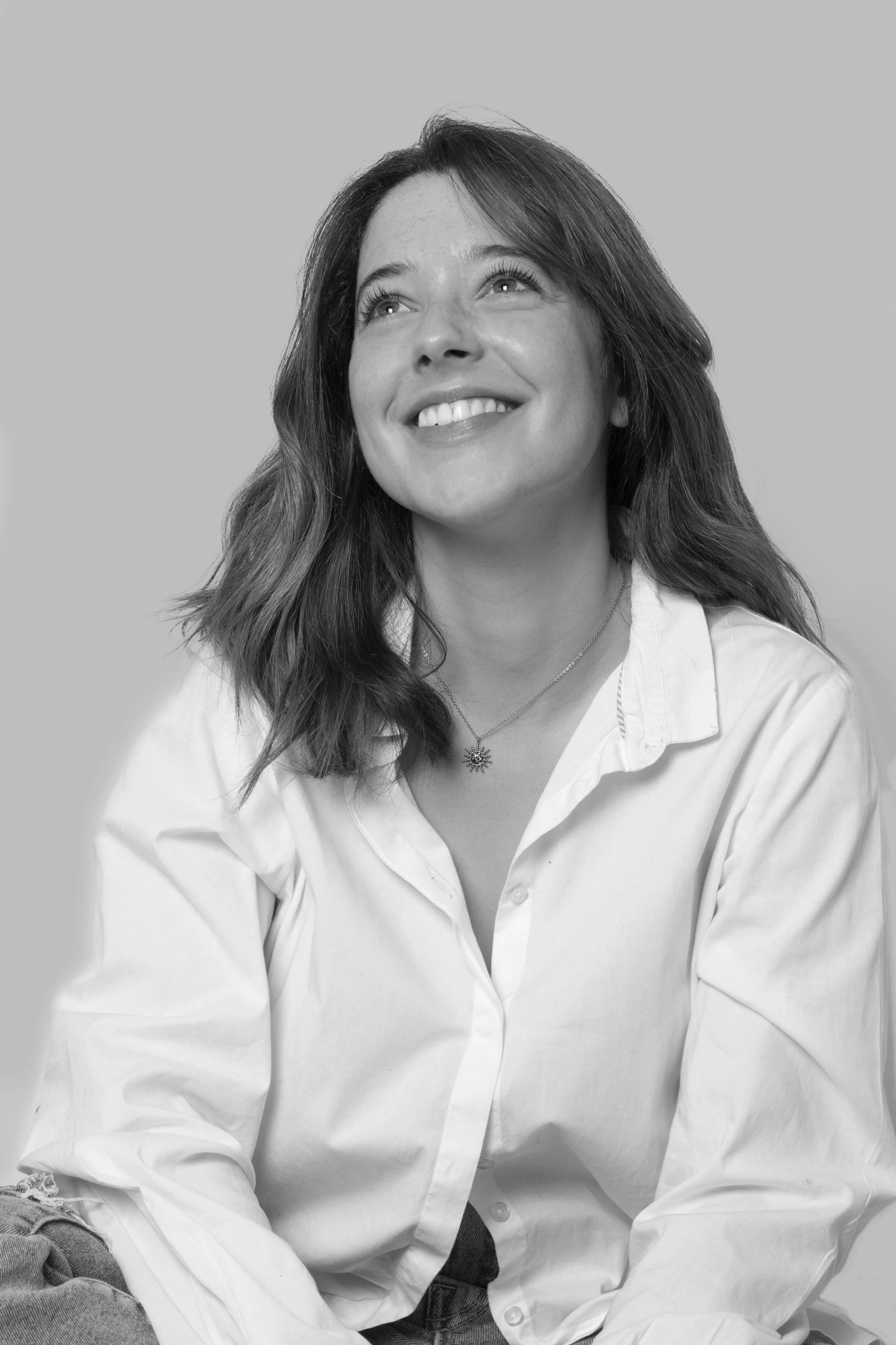 Portrait d'une femme souriante en chemise blanche, regardant vers le haut, dans un fond gris.