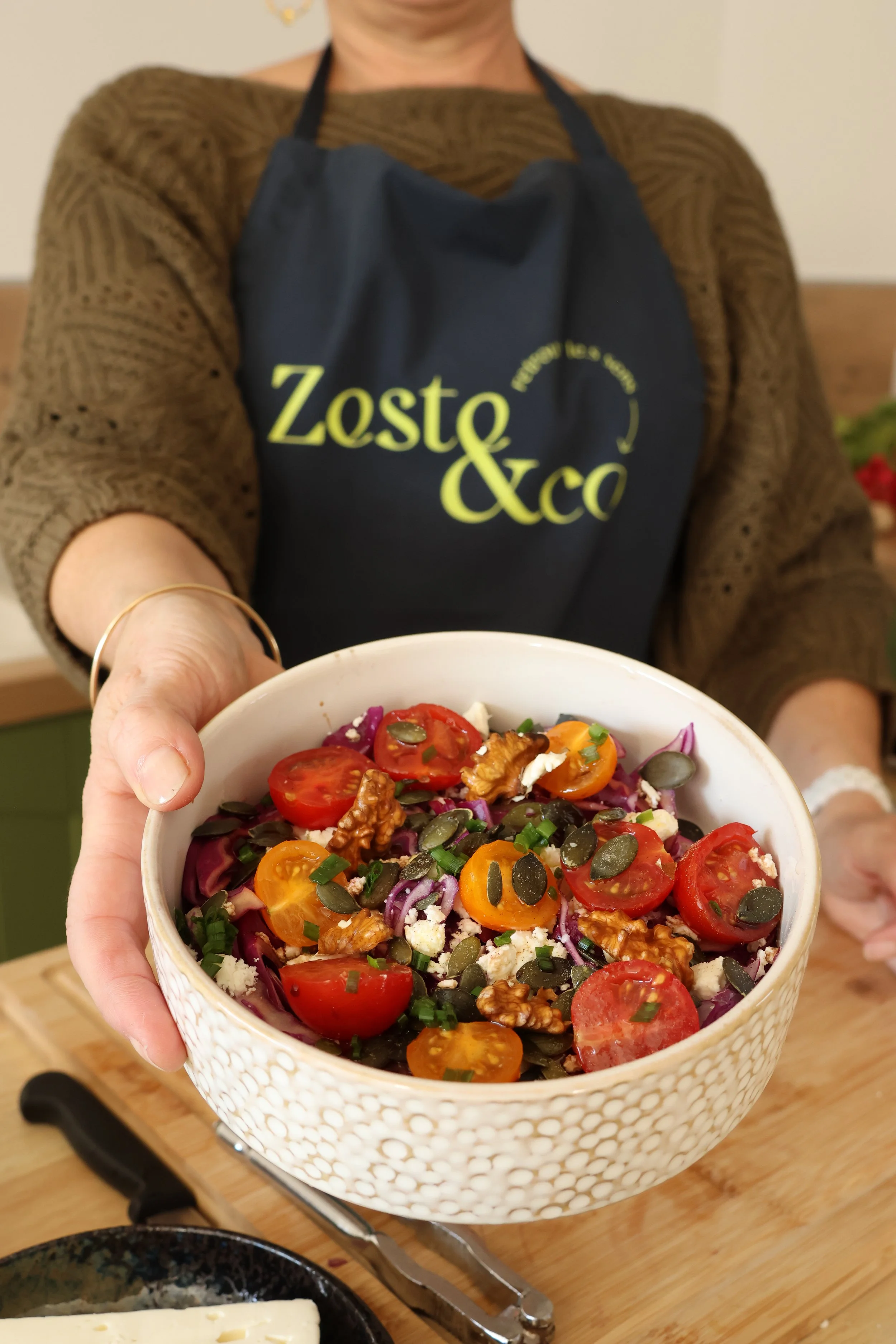 Une personne tenant un bol de salade colorée avec tomates cerises, noix, fromage, graines et oignons, en arrière-plan un plan de travail en bois.