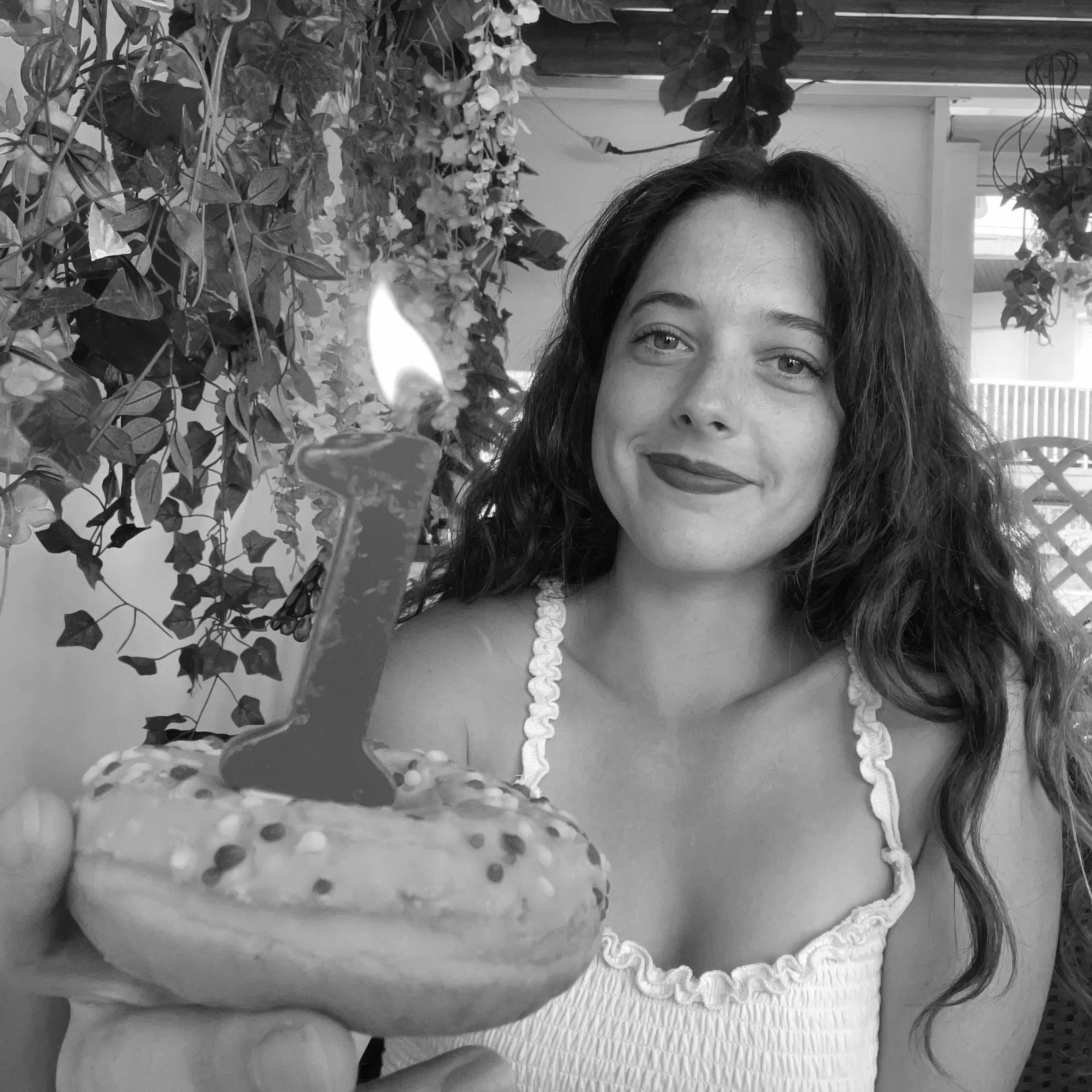 Une femme souriante tient un cupcake avec une bougie en forme de chiffre 1 allumée, en arrière-plan des plantes et un intérieur de maison.