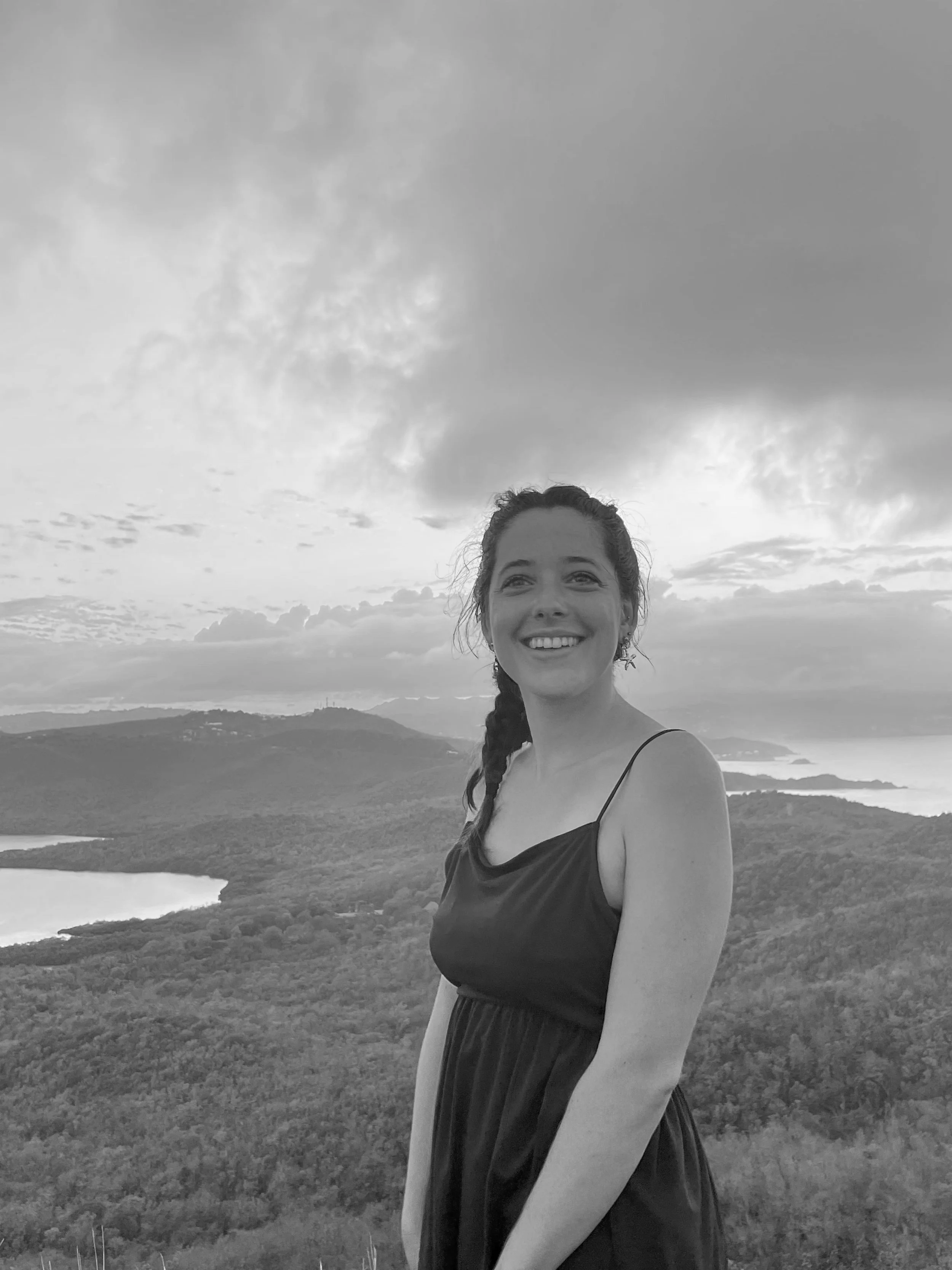 Une femme souriante portant une robe noire, avec un paysage vallonné, des lacs et la mer en arrière-plan, sous un ciel nuageux.