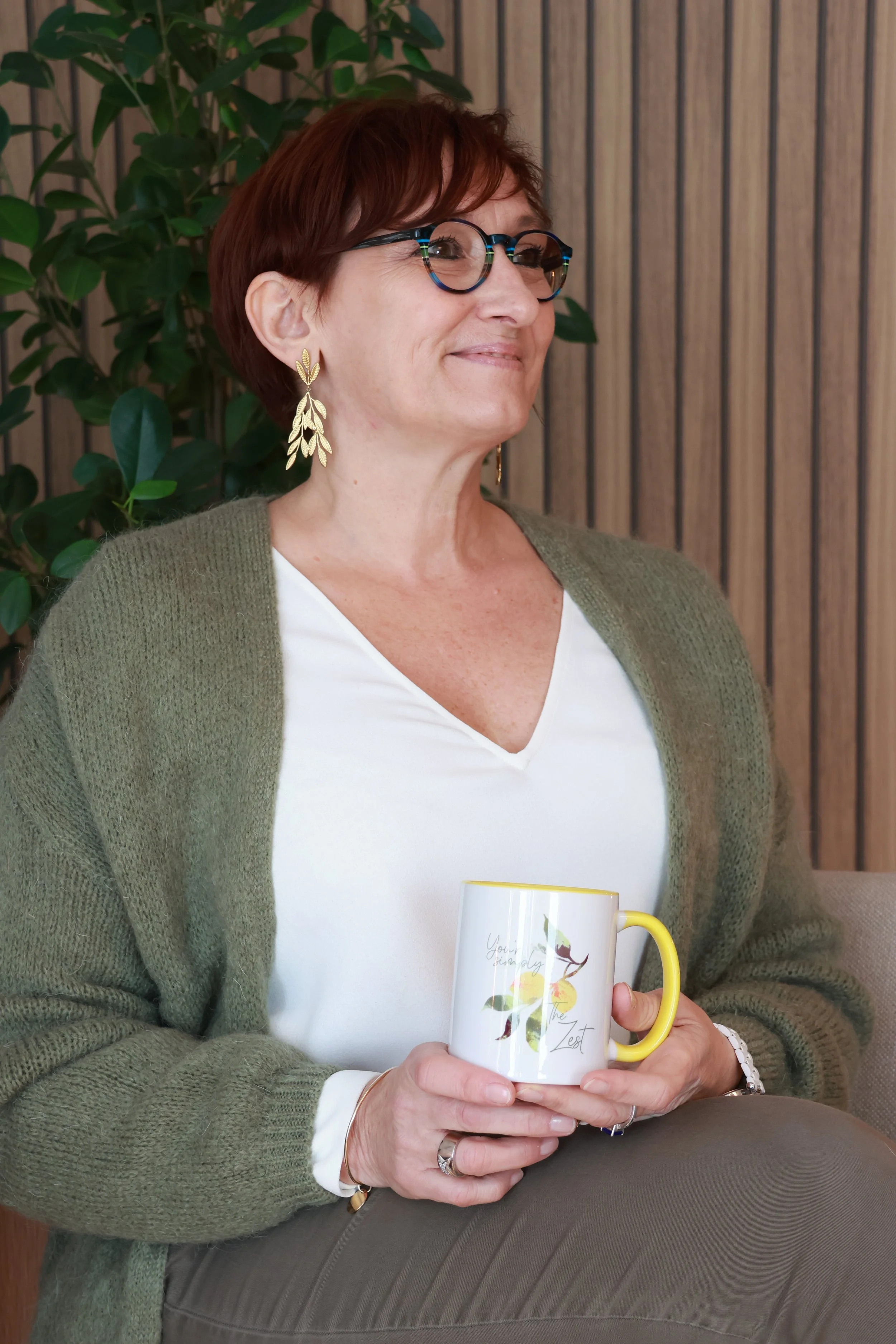 Femme assise tenant une tasse blanche avec un motif floral, portant des lunettes, des boucles d'oreilles en feuilles dorées, un pull vert et un chemisier blanc, avec un fond en bois et une plante verte.