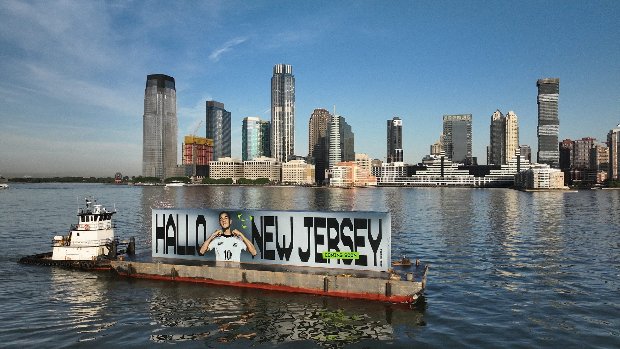 Tugboat pushing the 'Hallo New Jersey' billboard barge past the Jersey City skyline, Nike Football Jamal Musiala World Cup 2026