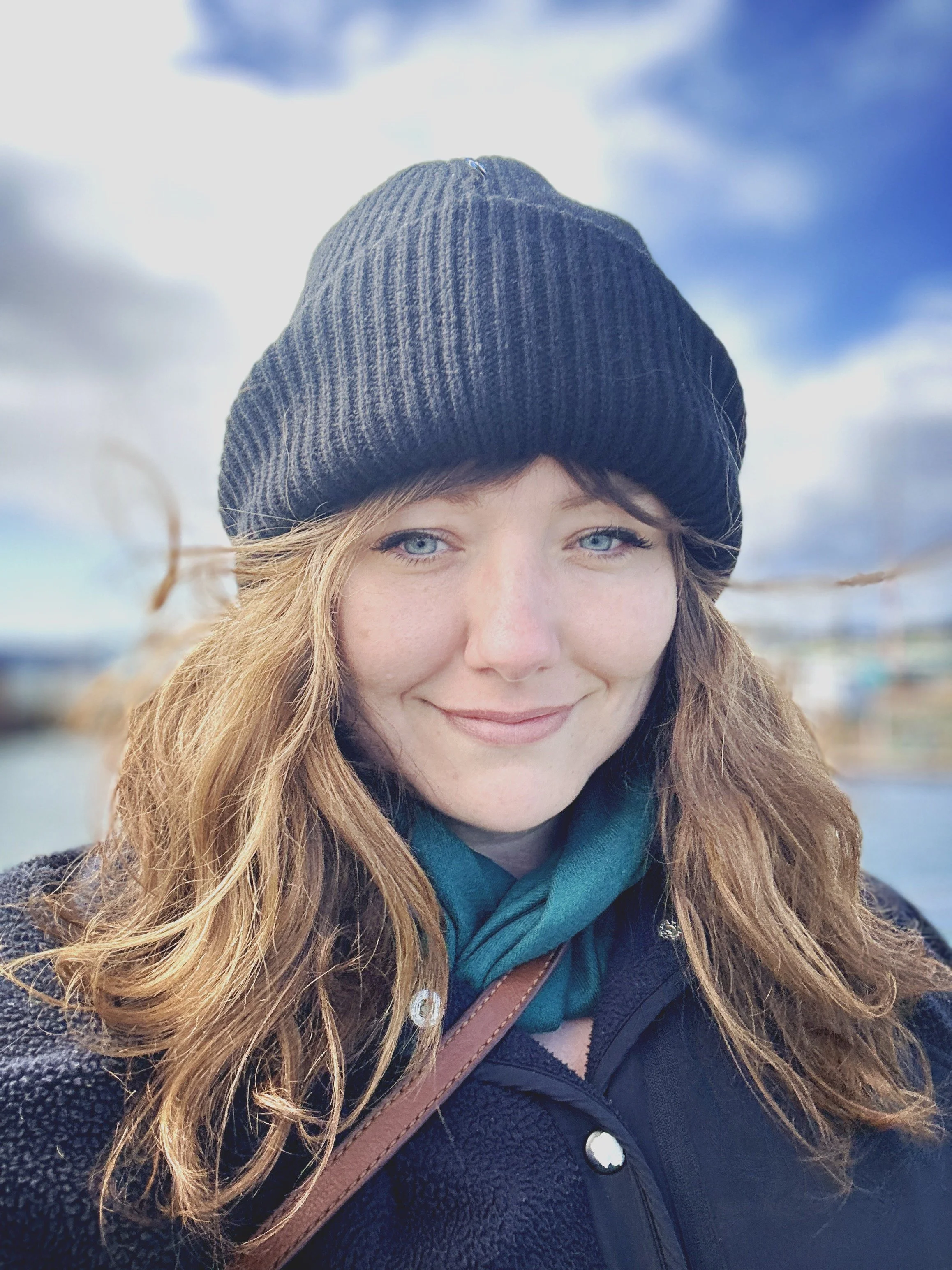 A woman with red hair, blue eyes, and freckles wearing a black beanie, teal scarf, black jacket, and carrying a brown purse.