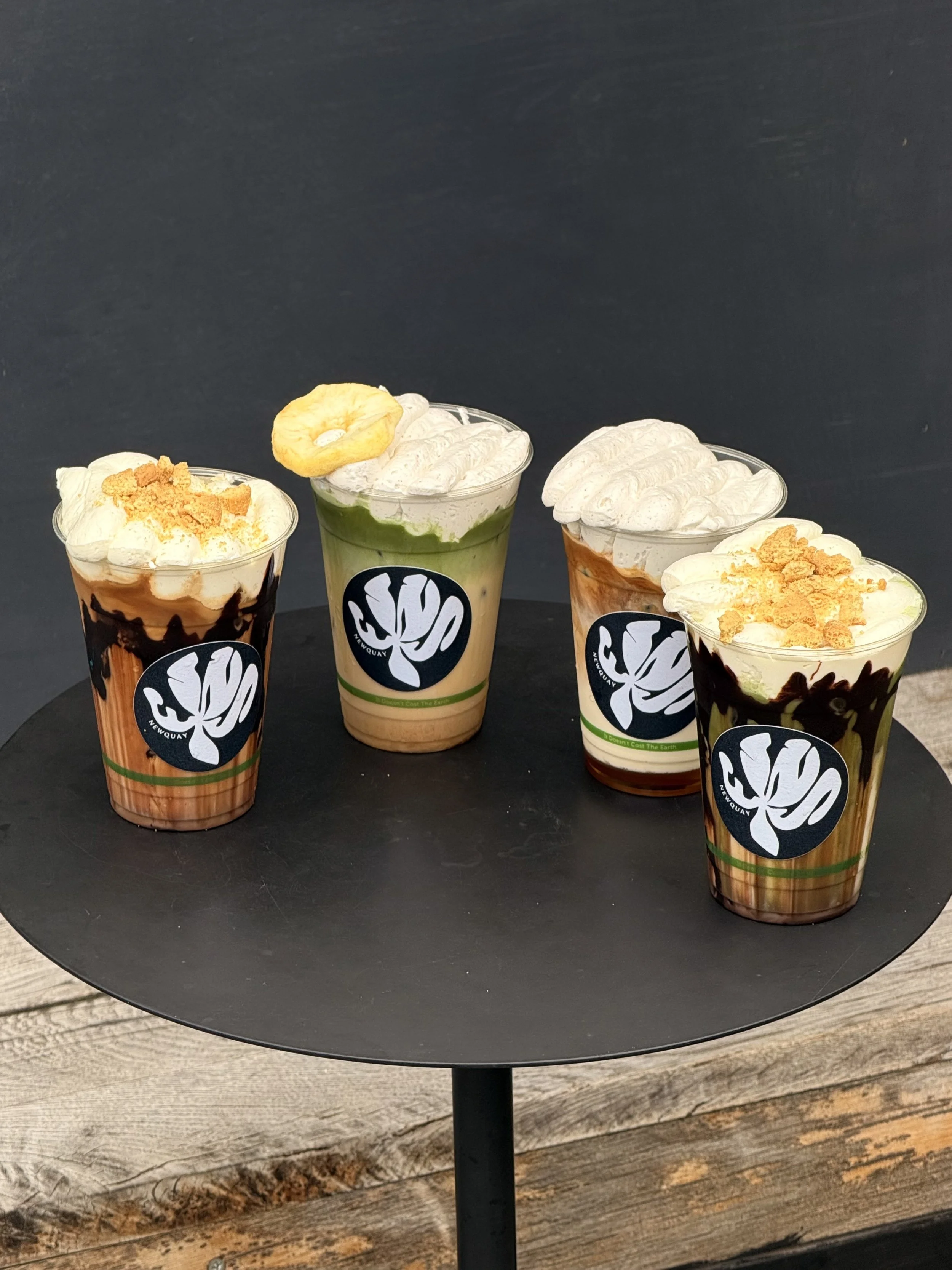 Four different ice cream drinks in clear plastic cups with whipped cream topping and various toppings, placed on a black round table.