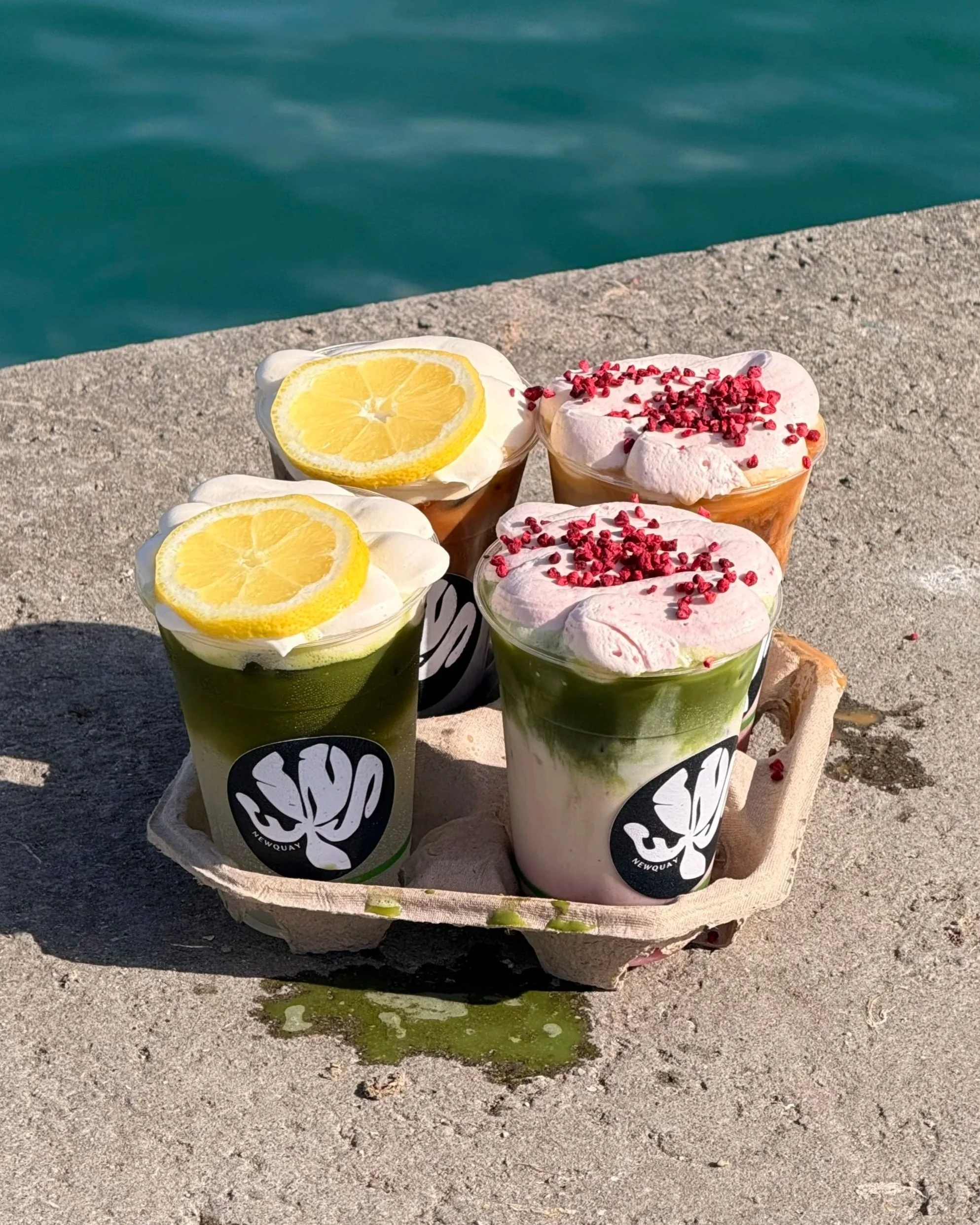 Four cups of layered matcha, vanilla, and red fruit desserts with whipped cream, lemon slices, and red berries, set on a stone surface near water.