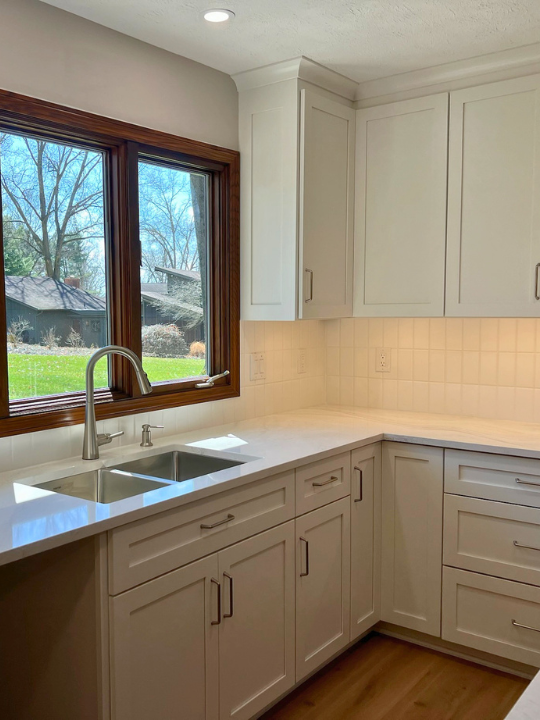 A kitchen with white cabinets, a white countertop, and a stainless steel sink with a modern faucet. There is a large window with wooden trim overlooking a backyard with trees and a house.