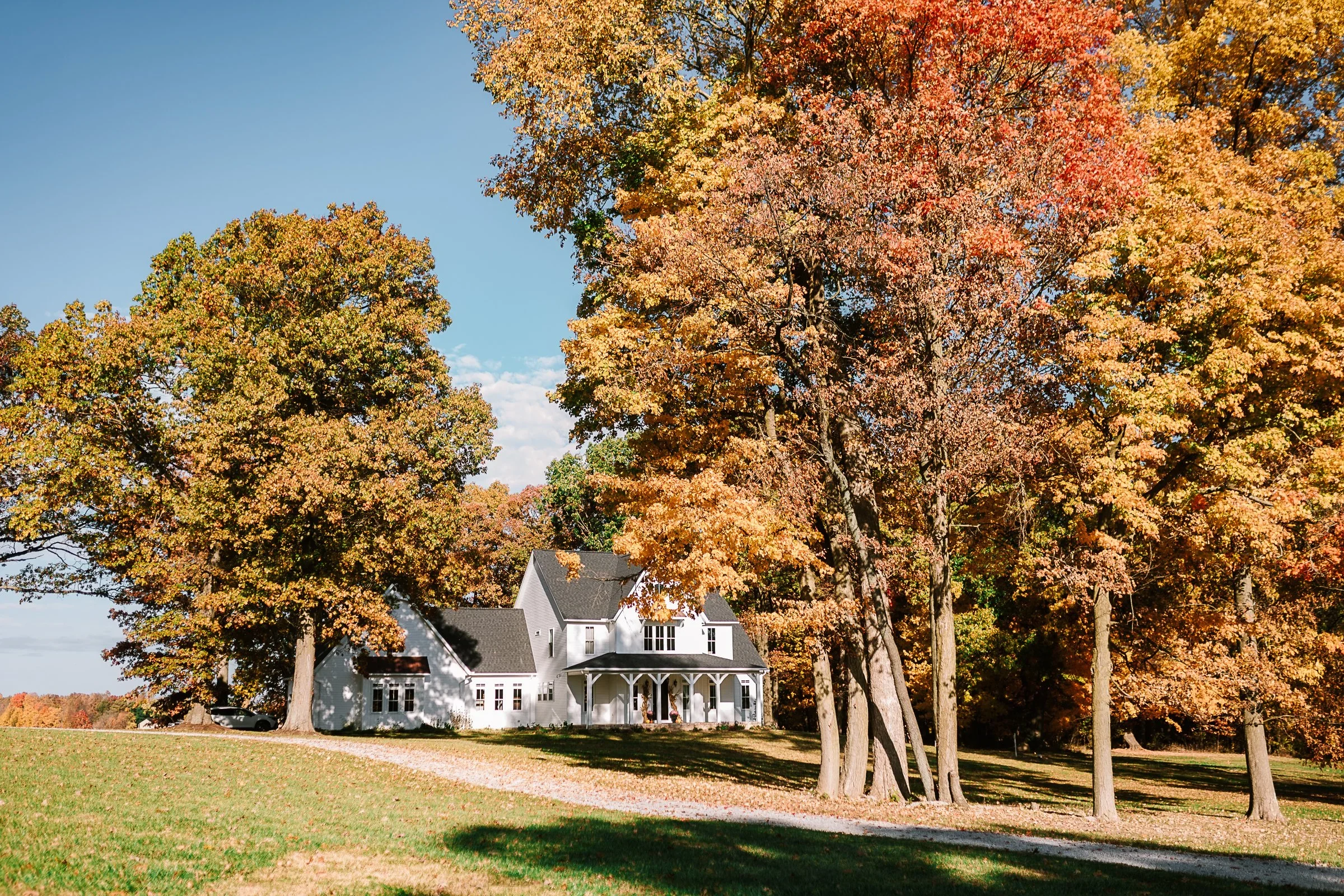 A white house with a dark roof surrounded by tall trees with colorful autumn leaves and a green lawn.