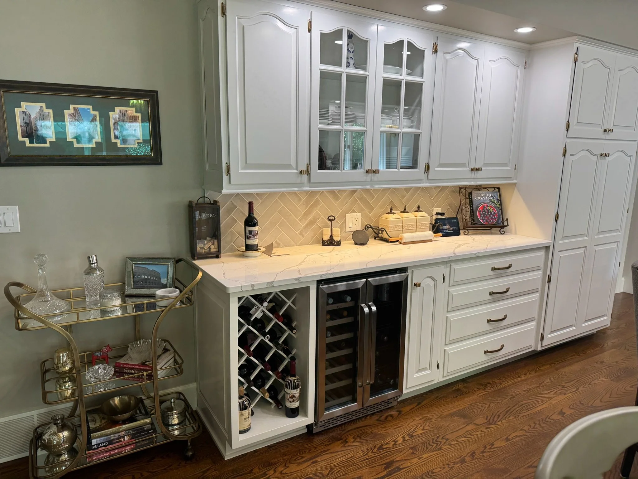 White kitchen cabinetry with glass-paneled doors, wine storage, and countertop decor including wine bottles, picture frames, and a cookbook, along with a small cart displaying glassware and books.