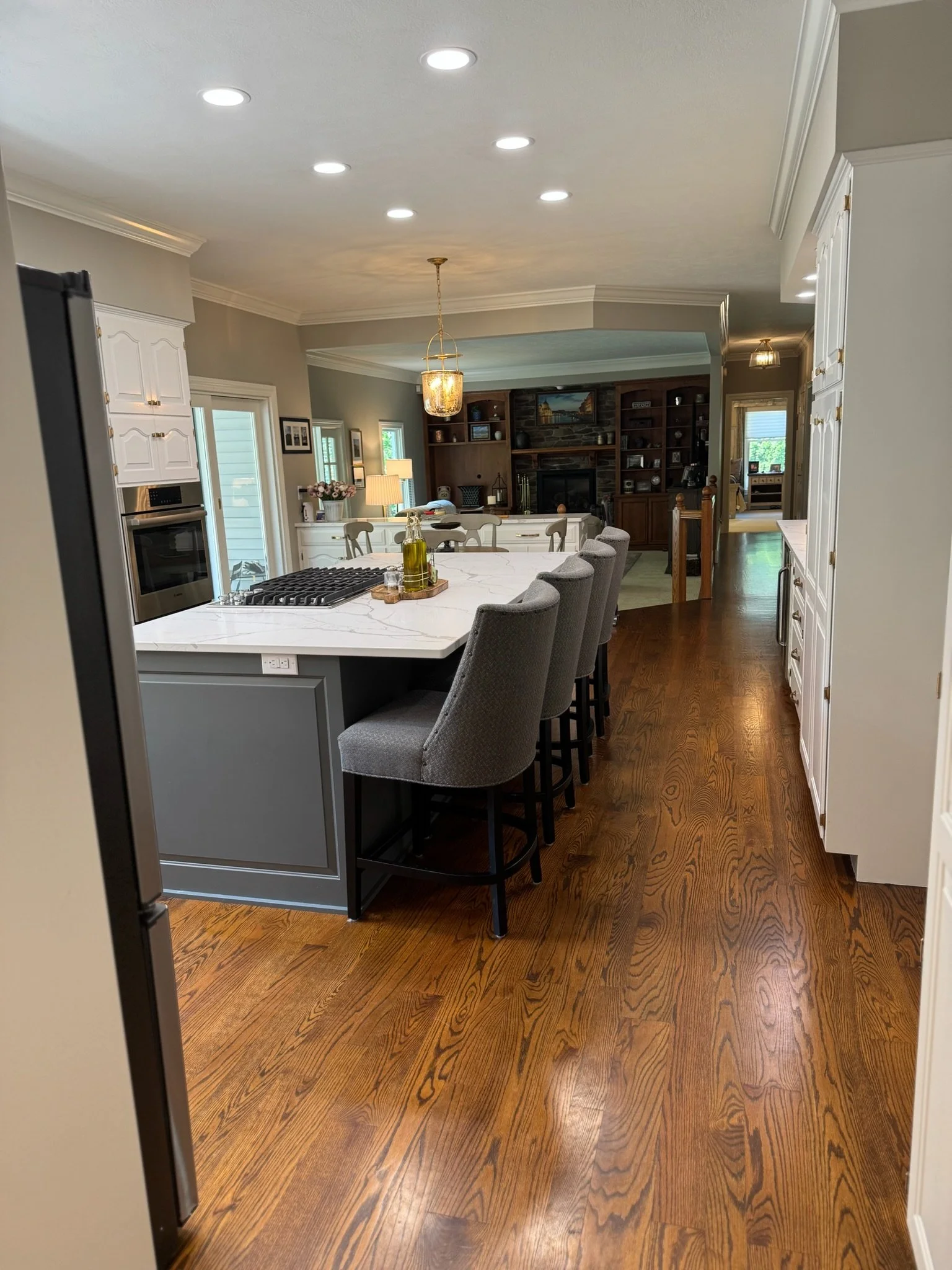 The image shows a modern kitchen and dining area with a white marble island, gray chairs, a wooden floor, and a fireplace with built-in shelves in the background, decorated with various items and a painting above the fireplace.