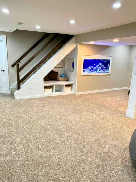 Basement with beige carpet, stairs with dark wood railing, built-in storage nook under stairs, and a wall-mounted aquarium with fish