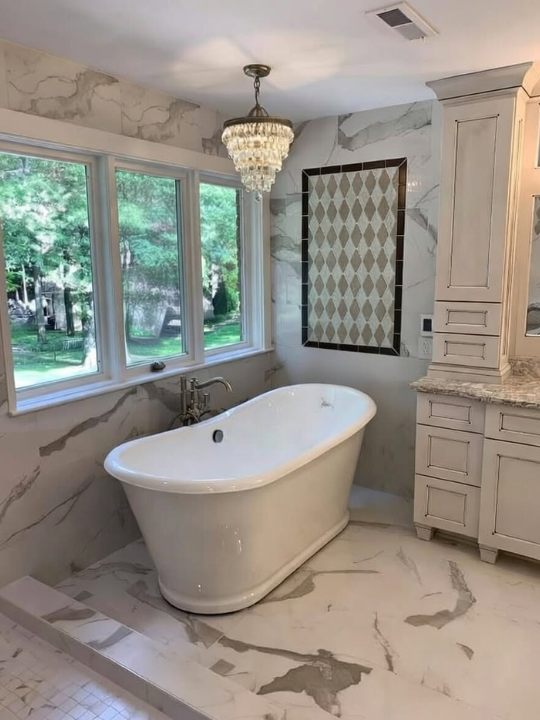 A luxurious bathroom featuring a freestanding bathtub beneath three large windows with a view of greenery outside. The bathroom has marble walls and flooring, a chandelier hanging from the ceiling, and a framed mirror on the wall. There is cabinetry with granite countertops on the right.