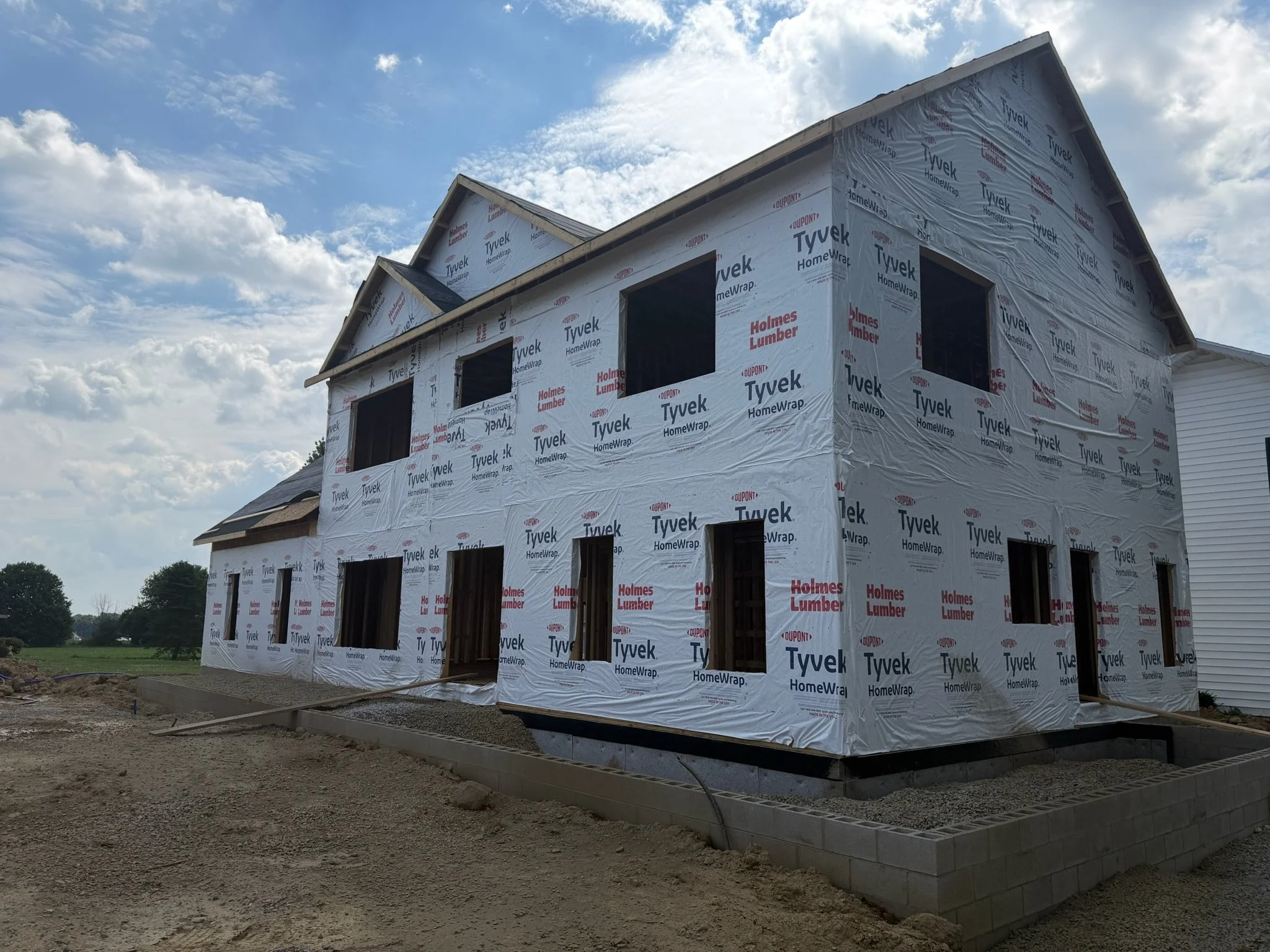 A two-story house under construction wrapped in white house wrap with branding, with a partly cloudy sky overhead.