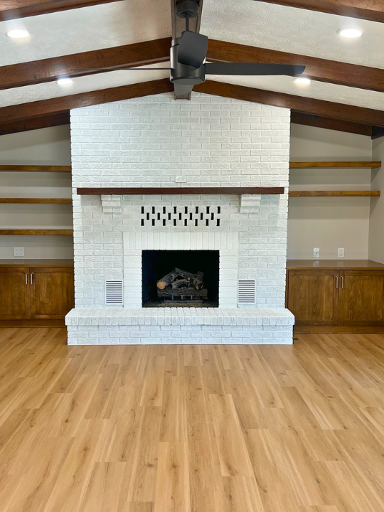 White brick fireplace with a wooden mantel, flanked by built-in wooden cabinets and open shelves on either side, in a room with wooden flooring and vaulted ceiling with dark wooden beams.