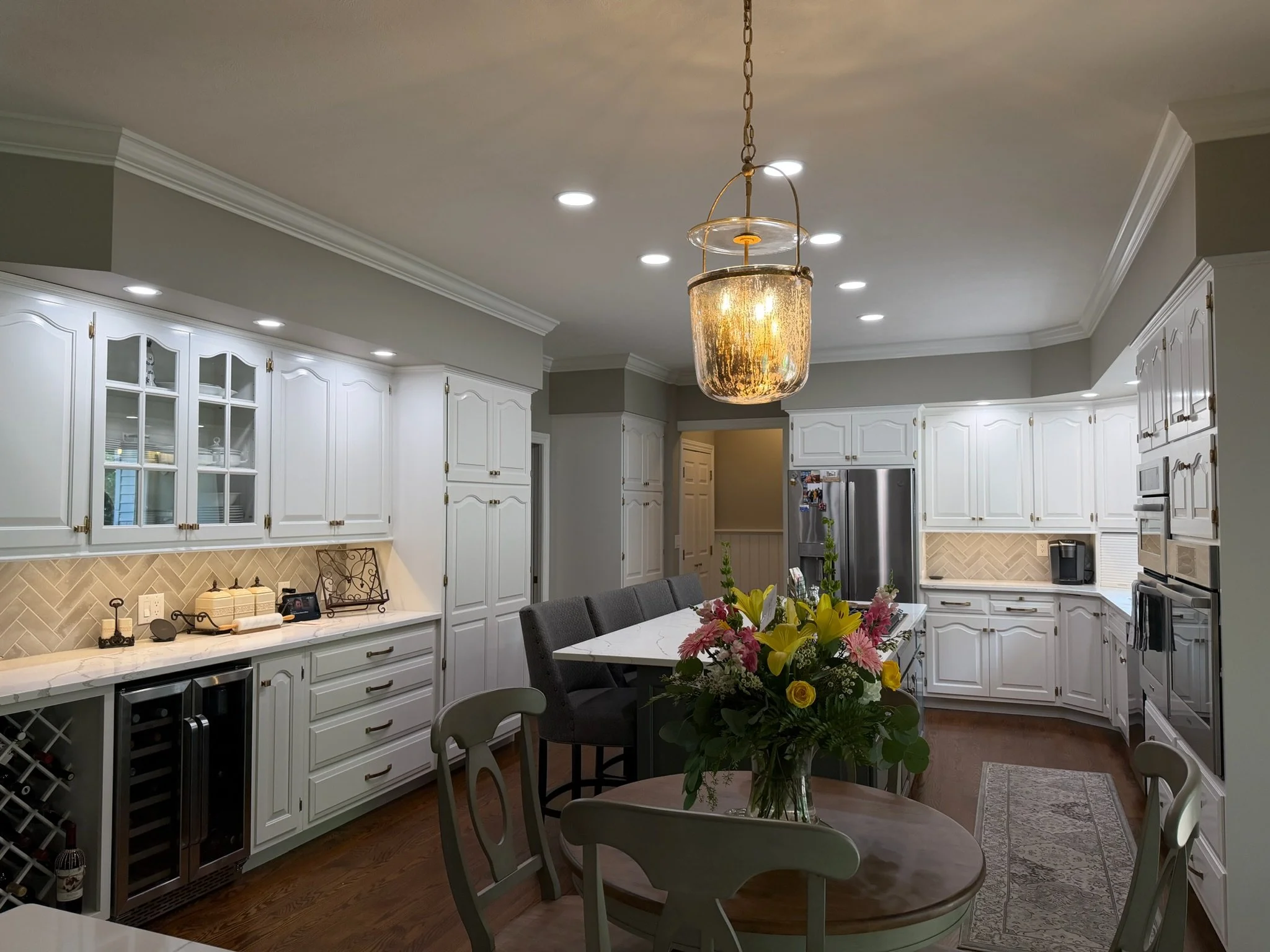 Kitchen with white cabinets, a central hanging light fixture, a wooden dining table with chairs, and a flower arrangement on the table.