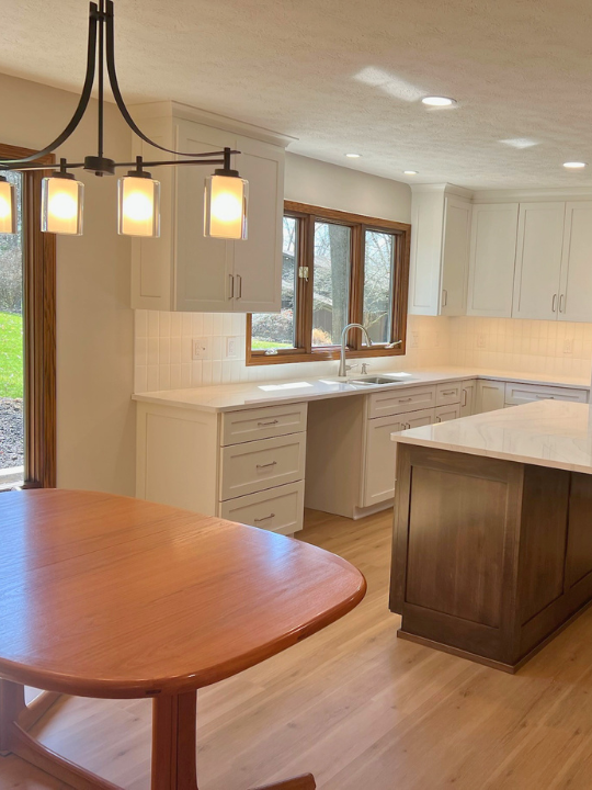 Modern kitchen with white cabinets, a large window, and a wooden island. There is a wooden dining table and a black pendant light fixture with five lights hanging from the ceiling.
