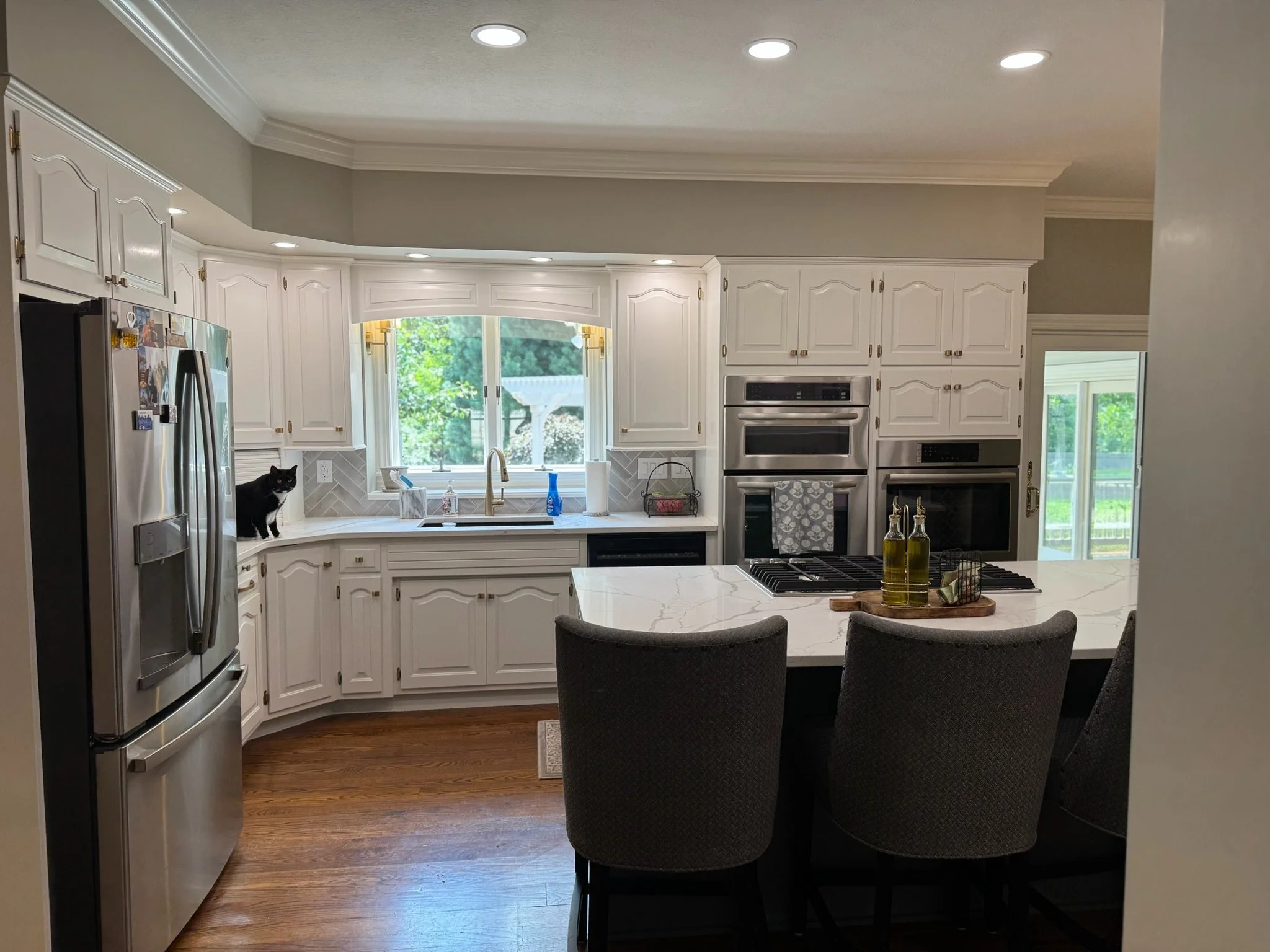 A modern kitchen with white cabinets, stainless steel appliances, a marble island, and black chairs. A black cat is sitting on the windowsill by the sink, and the view outside shows green trees.