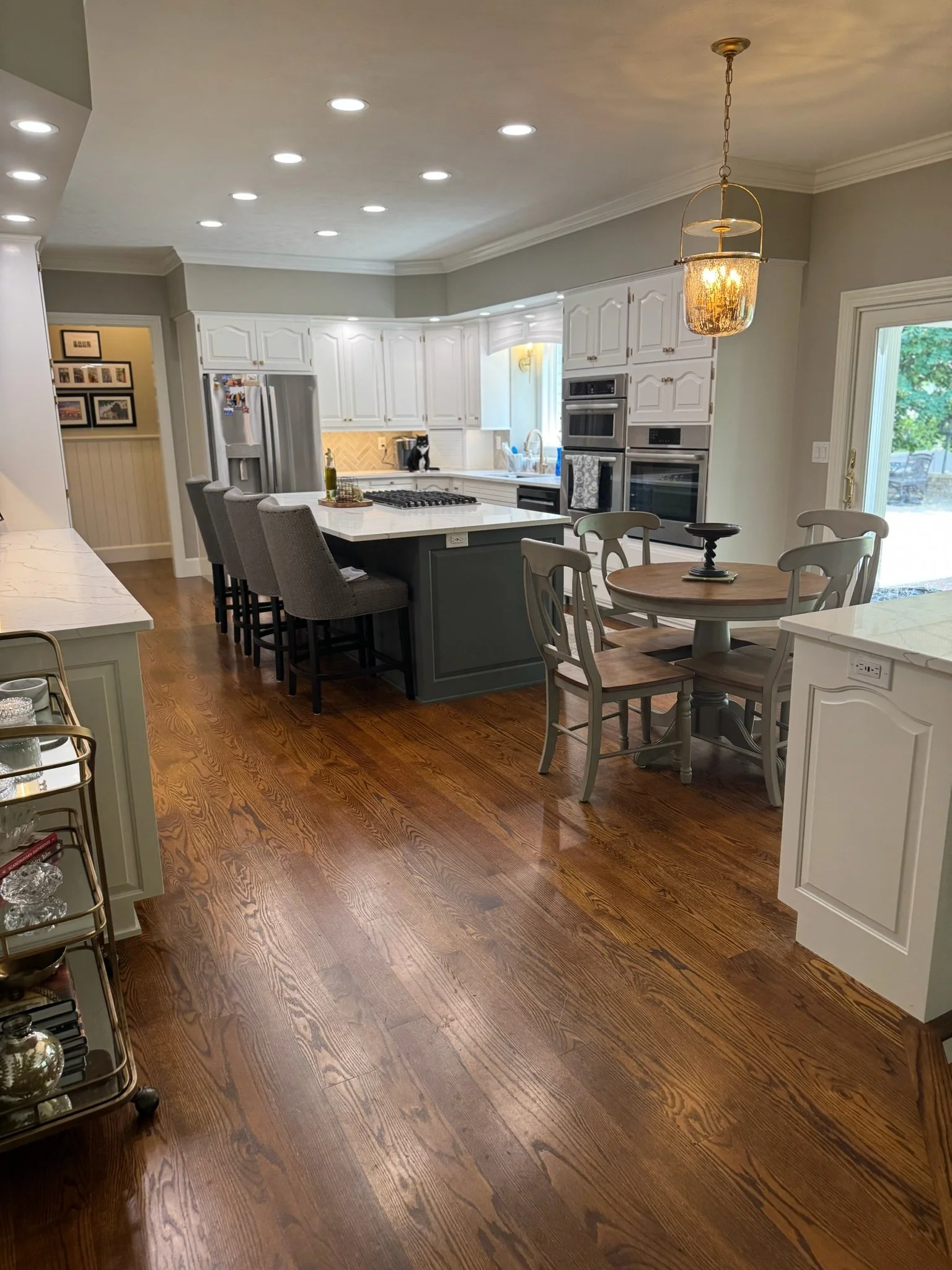 A spacious kitchen with hardwood floors, white cabinetry, and a kitchen island. There are multiple built-in ovens, a refrigerator, and a window with natural light. A black cat sits on the counter near the sink, and a decorative light fixture hangs from the ceiling. There are dining chairs around a round table and a sliding glass door leading outside.
