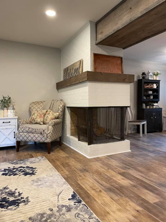 Living room corner with a cozy armchair, white side table, decorative pillows, wooden fireplace mantel, and hardwood flooring.