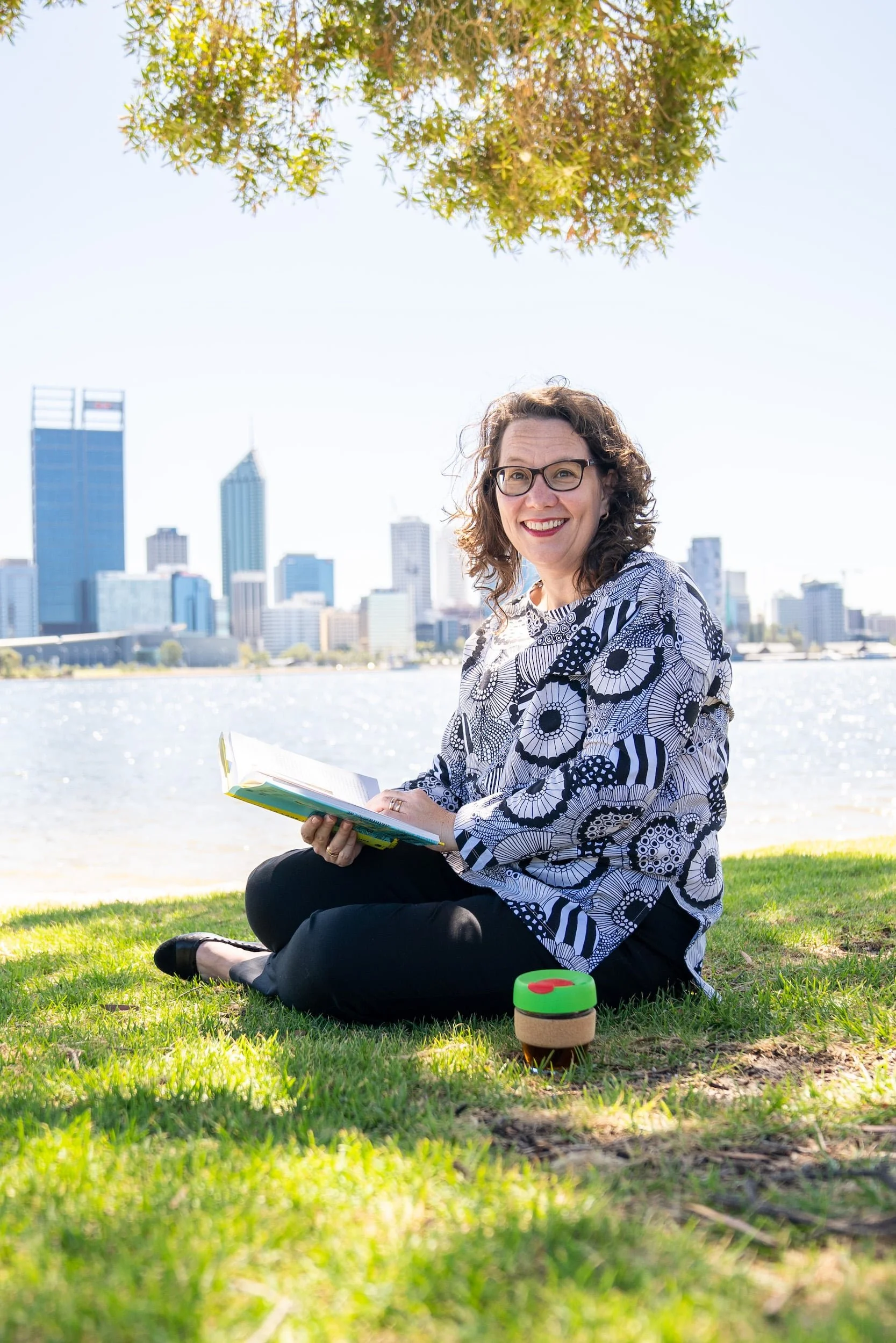 Rachael Bernstone reading on book on the grass, beside the Swan River, with the Perth skyline behind.