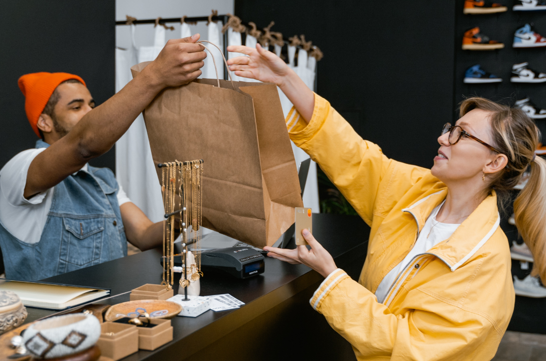 A woman with glasses in a yellow jacket paying for a brown paper bag at a boutique store while a man with an orange beanie hands her the bag. Jewelry and sneakers are visible in the background.