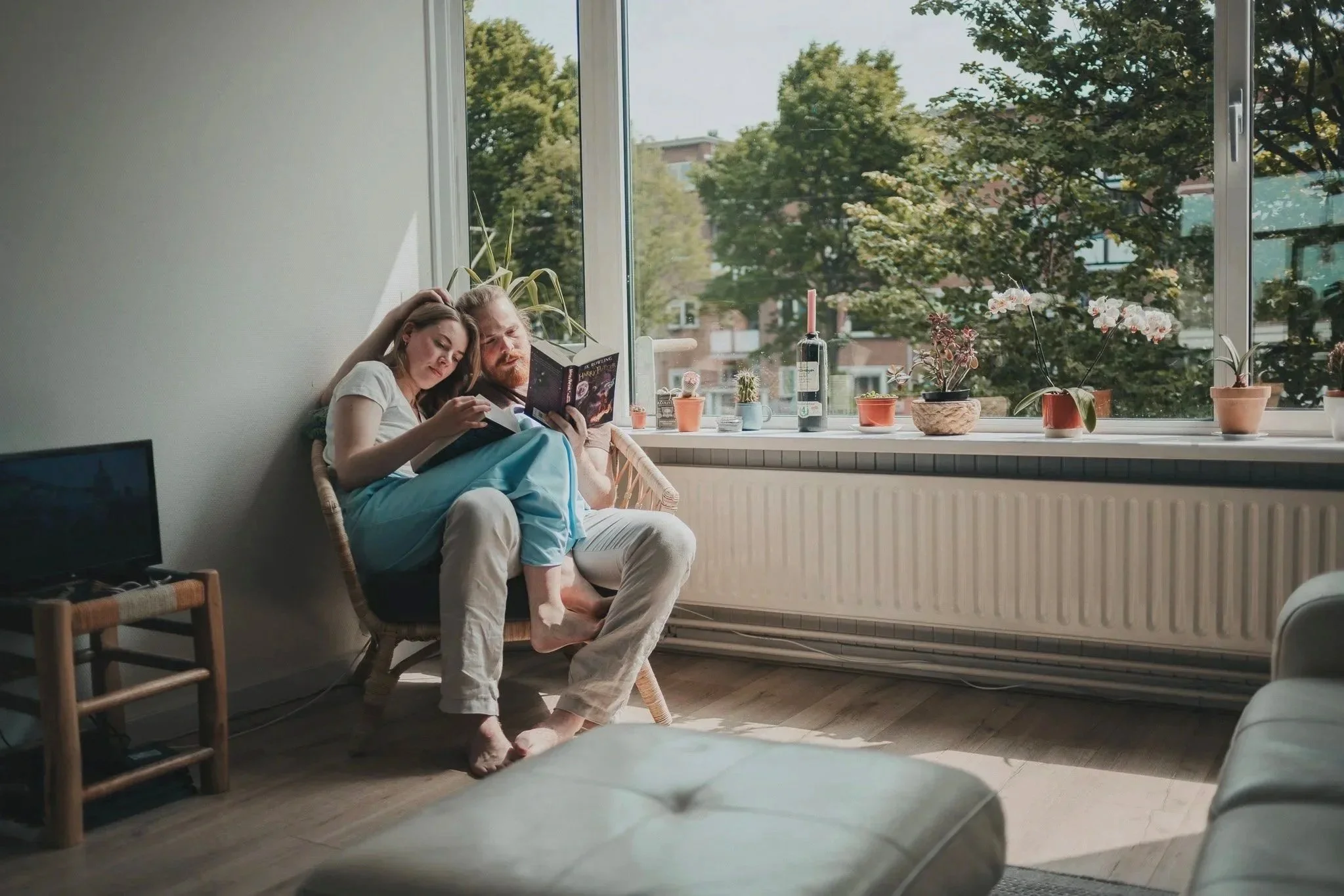 Twee mensen zitten samen in een woonkamer, leunend tegen de muur, terwijl ze een boek lezen. Het is zonnig buiten, te zien door het grote raam met planten op de vensterbank.