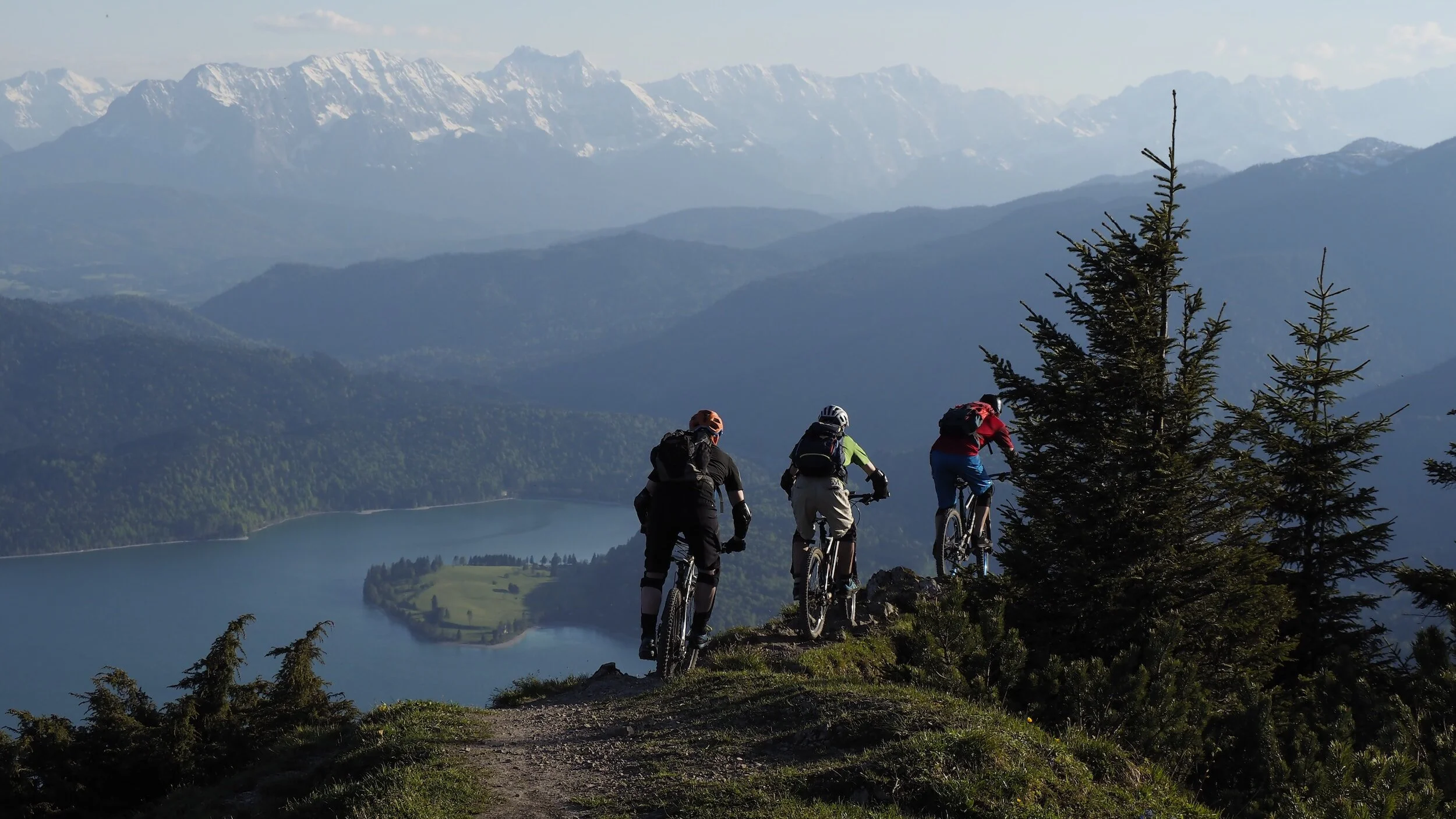 Ausblick von einem Berg auf einen See mit Bergen in der Weite und drei Mountainbike-Fahrer im Vordergrund. Rechtsanwalt Dr. Werner Mecenovic