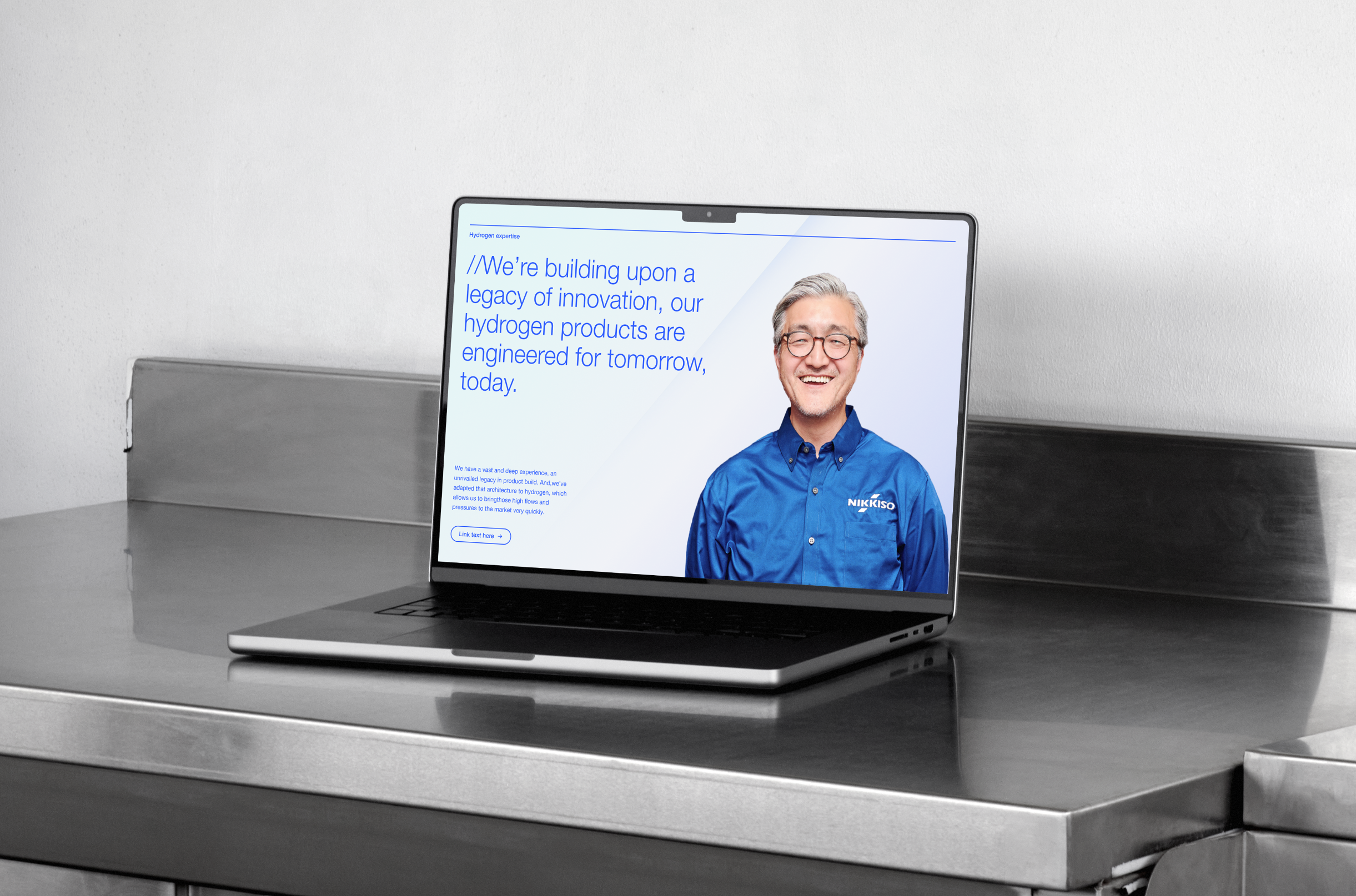 A laptop on a metallic surface displaying a webpage with a smiling man in glasses and a blue dress shirt with Nikkiso logo, alongside blue text about hydrogen products.