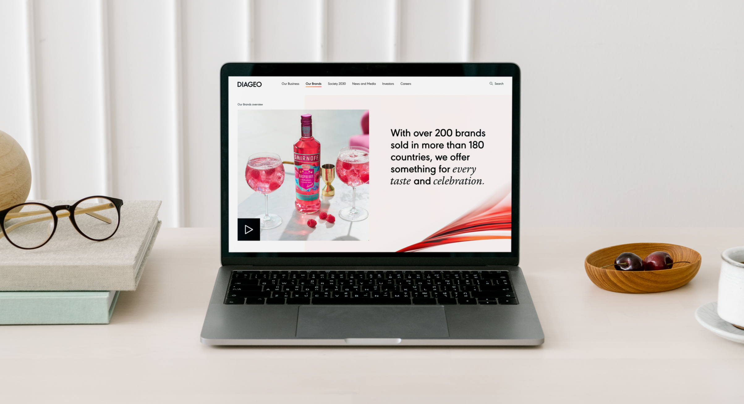 Open laptop displaying a website about brands, with an image of pink beverages with raspberries and a vodka bottle. On the left, there are glasses and a box of glasses on a notebook. To the right, a wooden bowl with cherries and a cup of coffee.