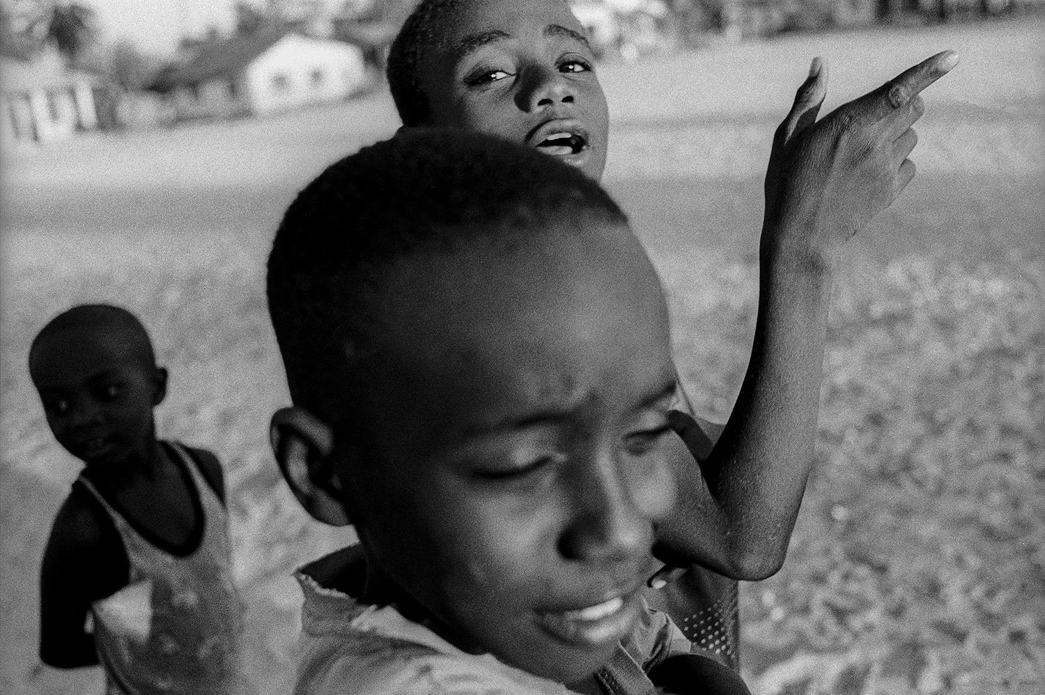 Black and white photograph of four children outdoors, with three girls in the foreground and a girl in the background, in a rural area with houses in the distance.