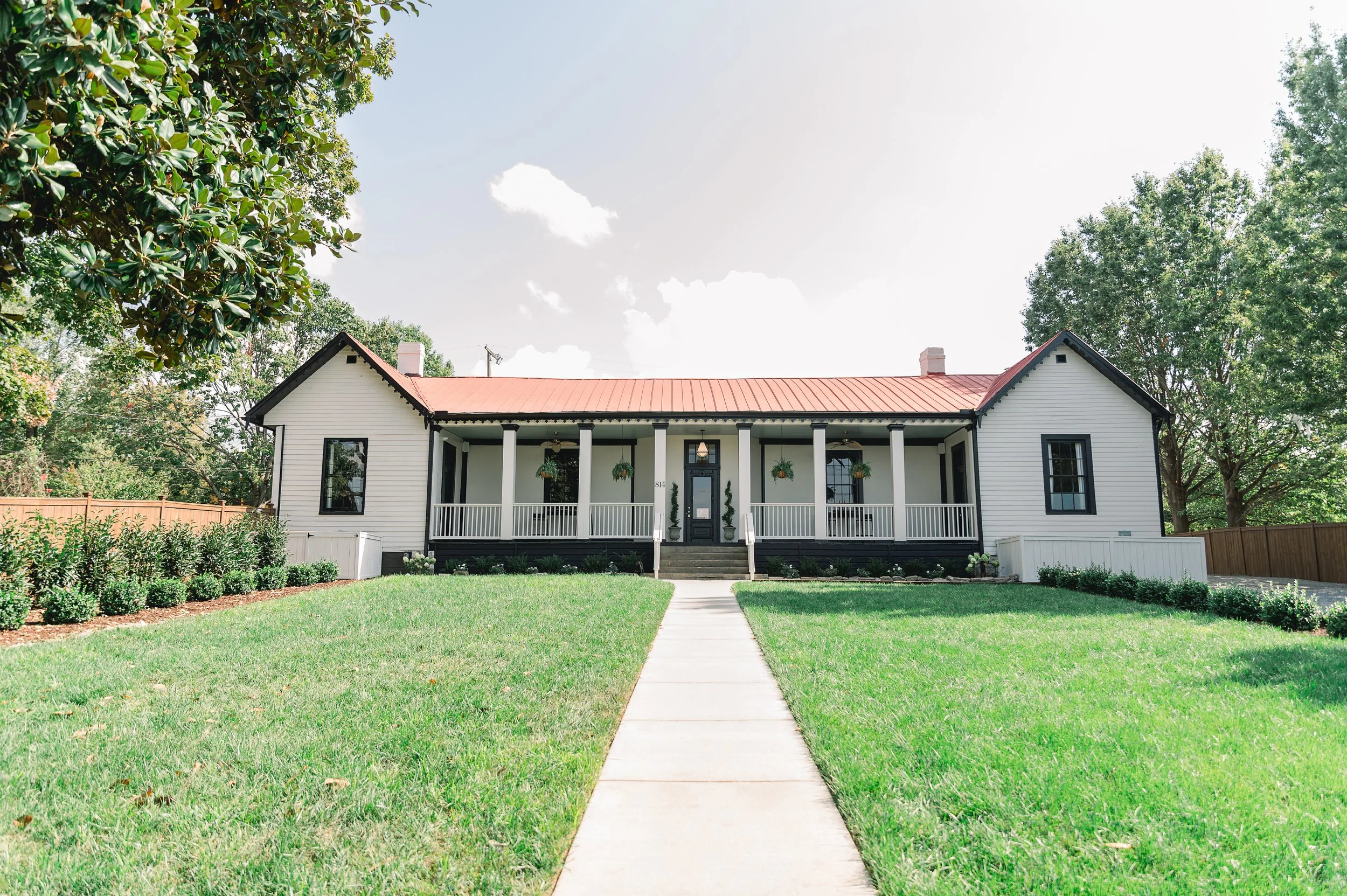 Front view of an East Nashville wedding venue, white Victorian house with black window frames, a red roof, and a large front porch with white railings, surrounded by green grass and trees.