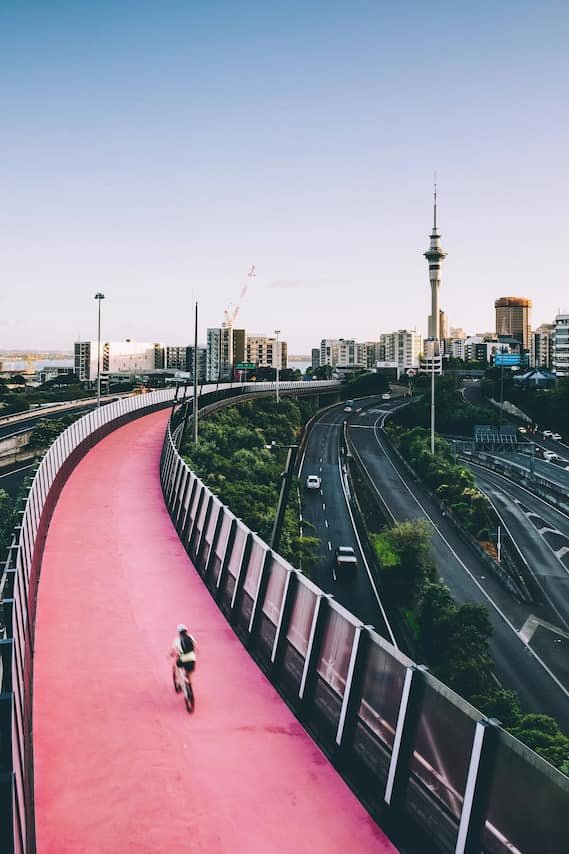 A person riding a bike on a pink elevated cycling path above city streets with skyscrapers and a prominent tower in the background.