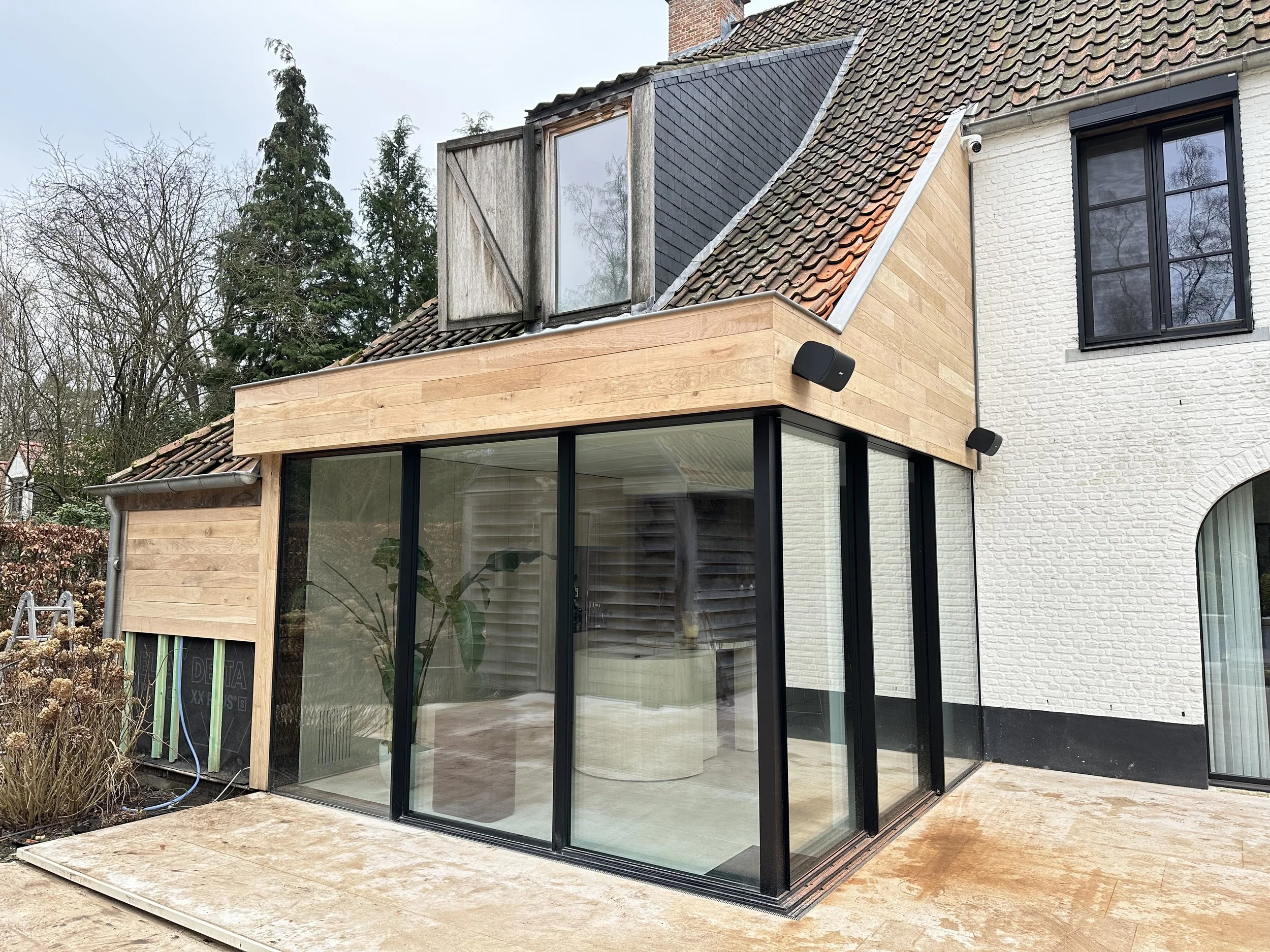 Moderne uitbouw in Geel met grote glazen ramen en een houten gevel, waarbij dakwerken worden uitgevvoerd