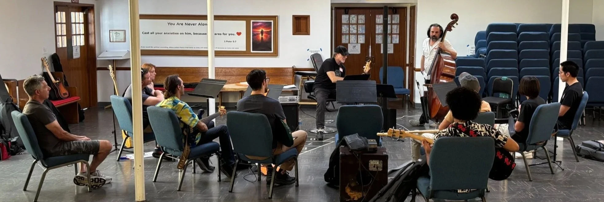 Group of musicians practicing in a church or community room, with some playing guitars, piano, and double bass, and one person conducting or giving instructions. Audience chairs are visible, some with stacked seating in the background, and a wall decorated with posters and a religious quote.