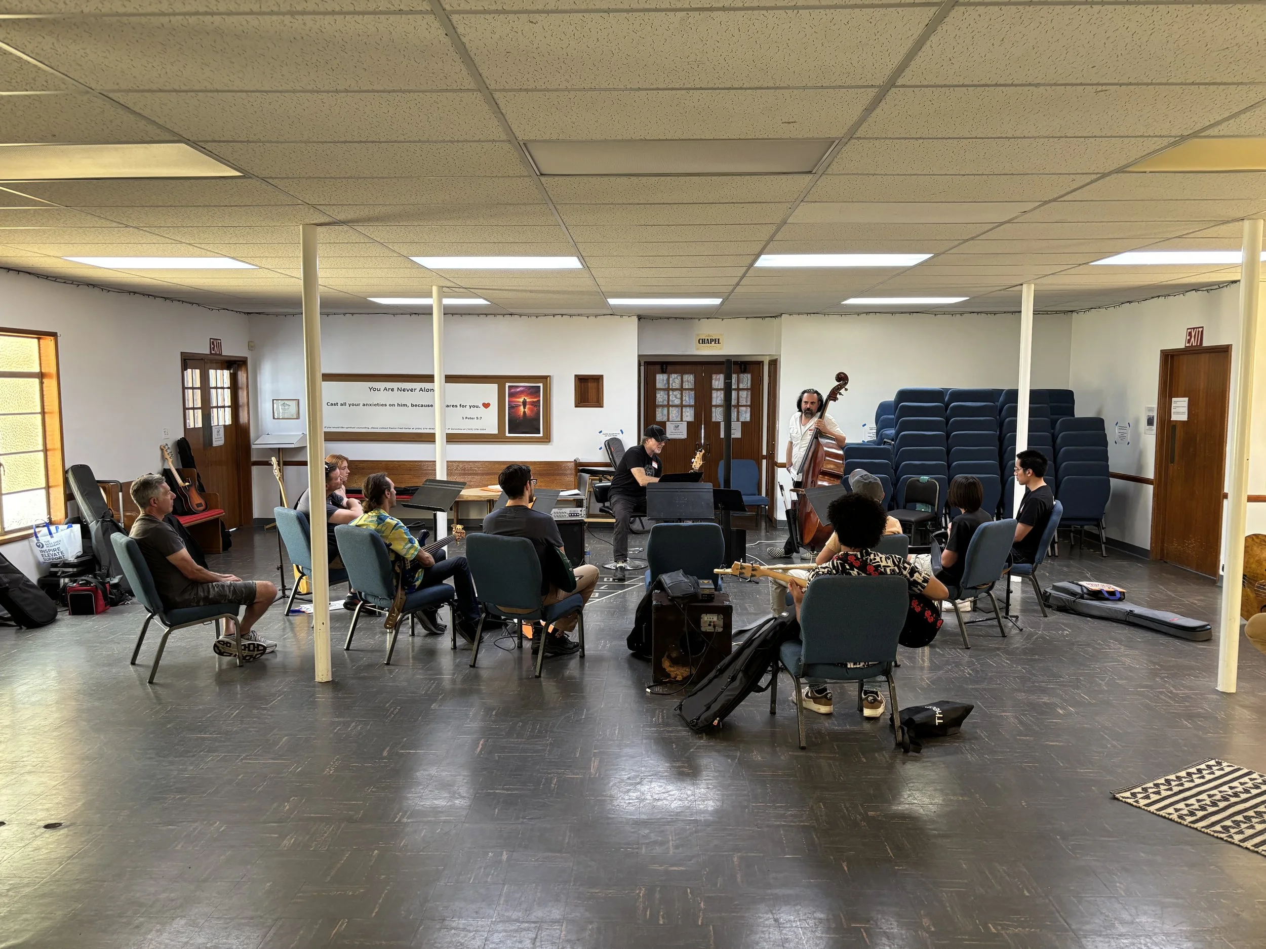 A group of people in a room, some sitting and some playing musical instruments, with a man standing and playing a double bass. There are chairs, guitars, and a mural with religious and inspirational text on the wall.