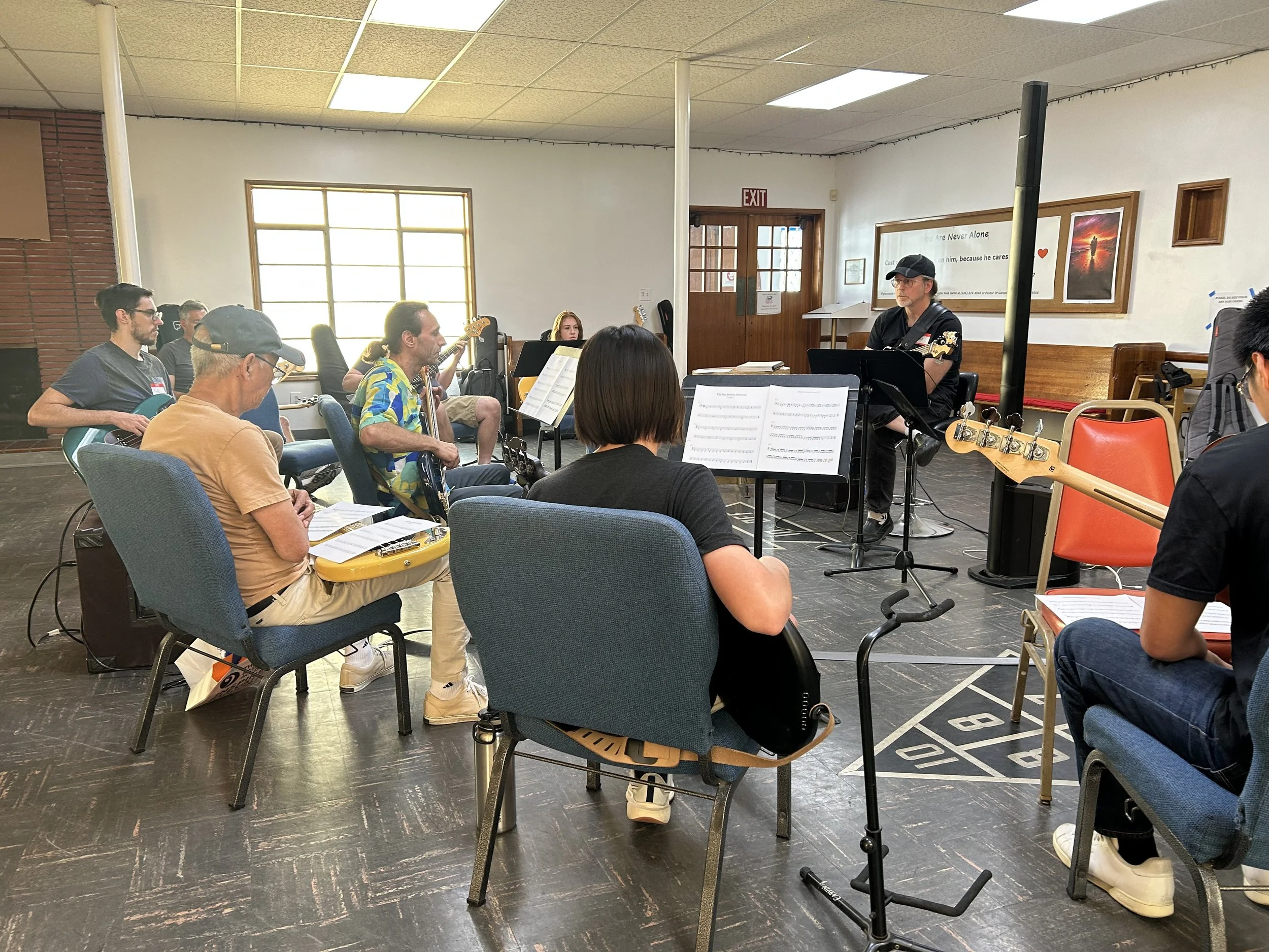 A group of people in a music rehearsal room, seated and holding guitars, with sheet music on stands in front of them. A man on the right appears to be leading or instructing. The room has large windows, wooden accents, and a casual atmosphere.