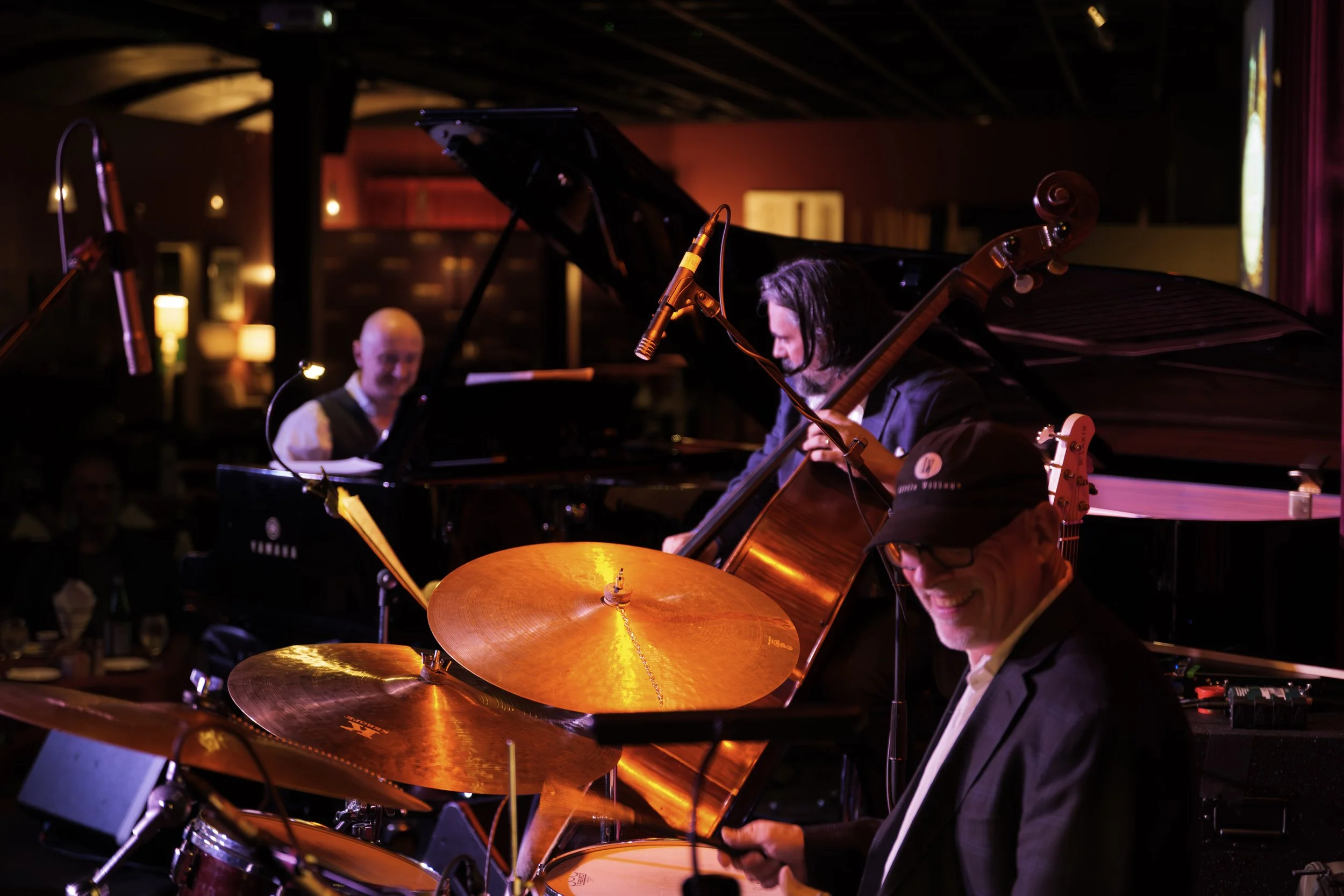 Jazz musicians playing instruments on stage in a dimly lit venue, including a pianist, a double bassist, and a drummer.