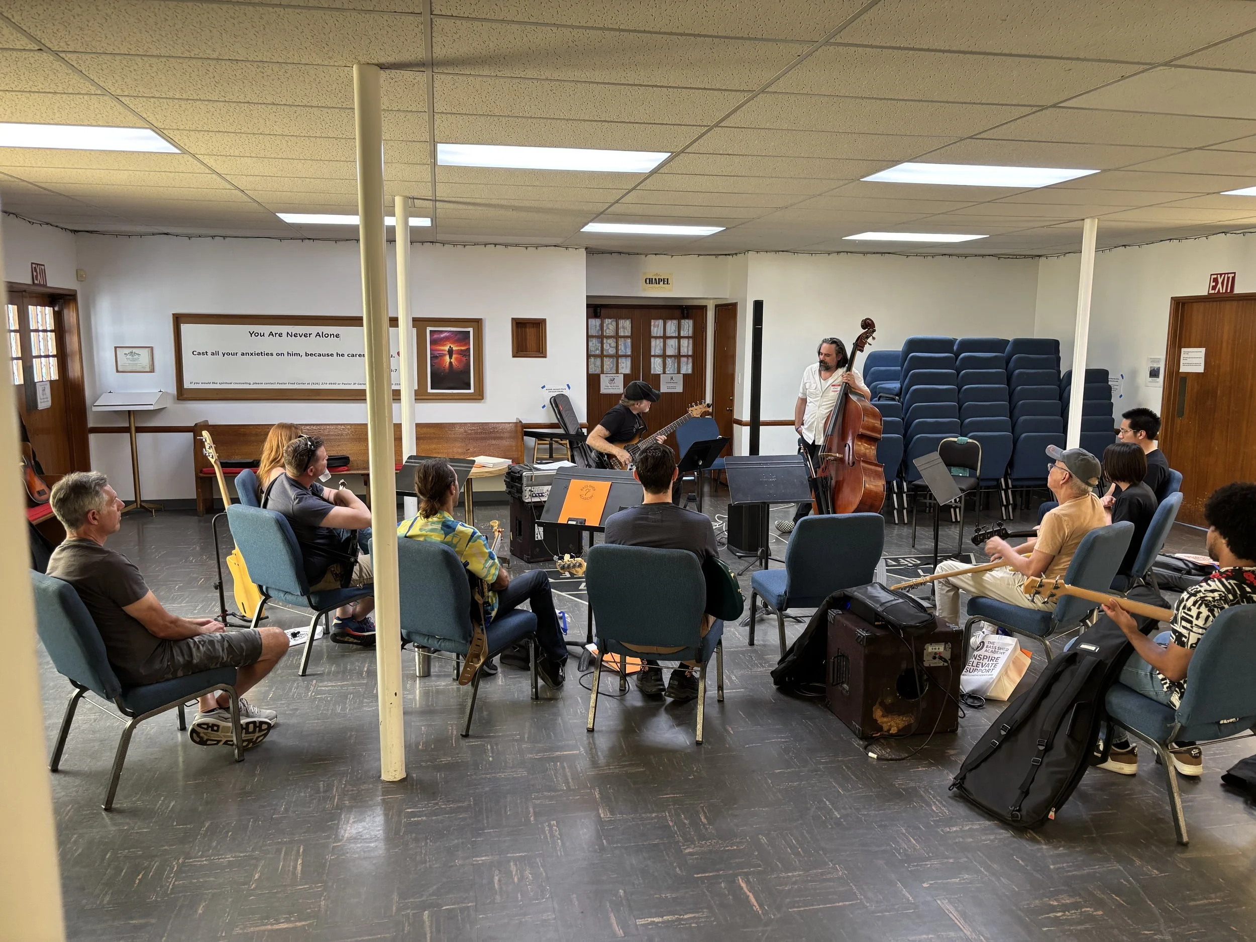 A group of people seated in chairs watch a live music performance with a man playing a double bass and another person playing a guitar in a community room.