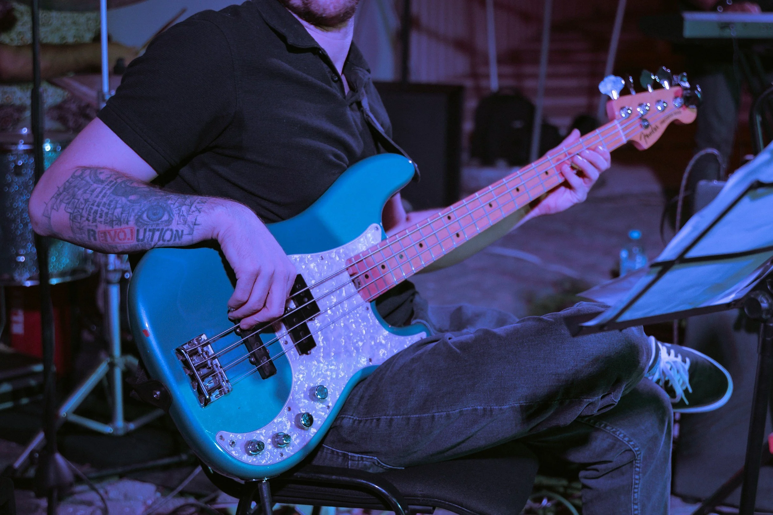 A person playing a blue electric bass guitar, sitting on a chair with one leg crossed over the other, surrounded by musical equipment and a music stand in an indoor or stage setting.