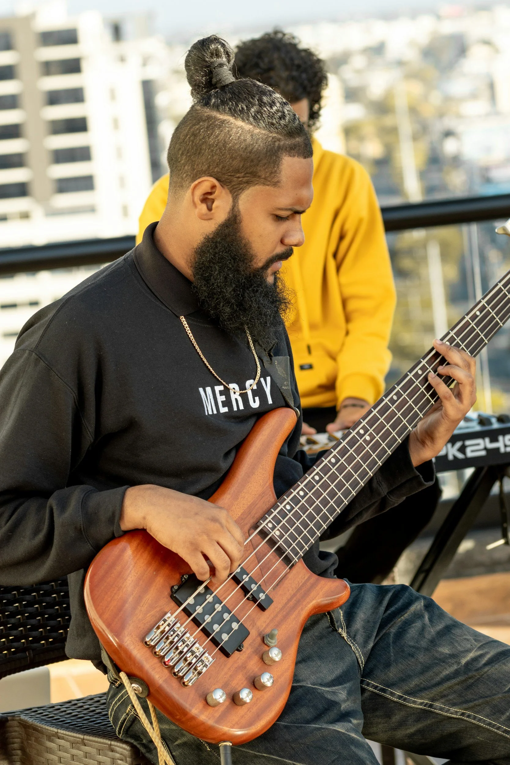 A man with a beard and tied-up hair playing an electric bass guitar, and another person in a yellow hoodie playing a keyboard in the background on a rooftop with city buildings.