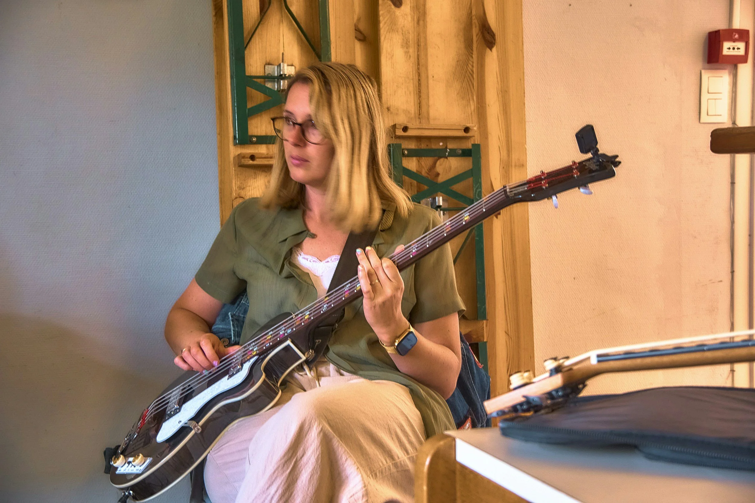 A woman with shoulder-length blonde hair, wearing glasses and a green shirt, is playing an electric guitar. She is sitting indoors, and there are wooden shelves and a fire alarm on the wall behind her.